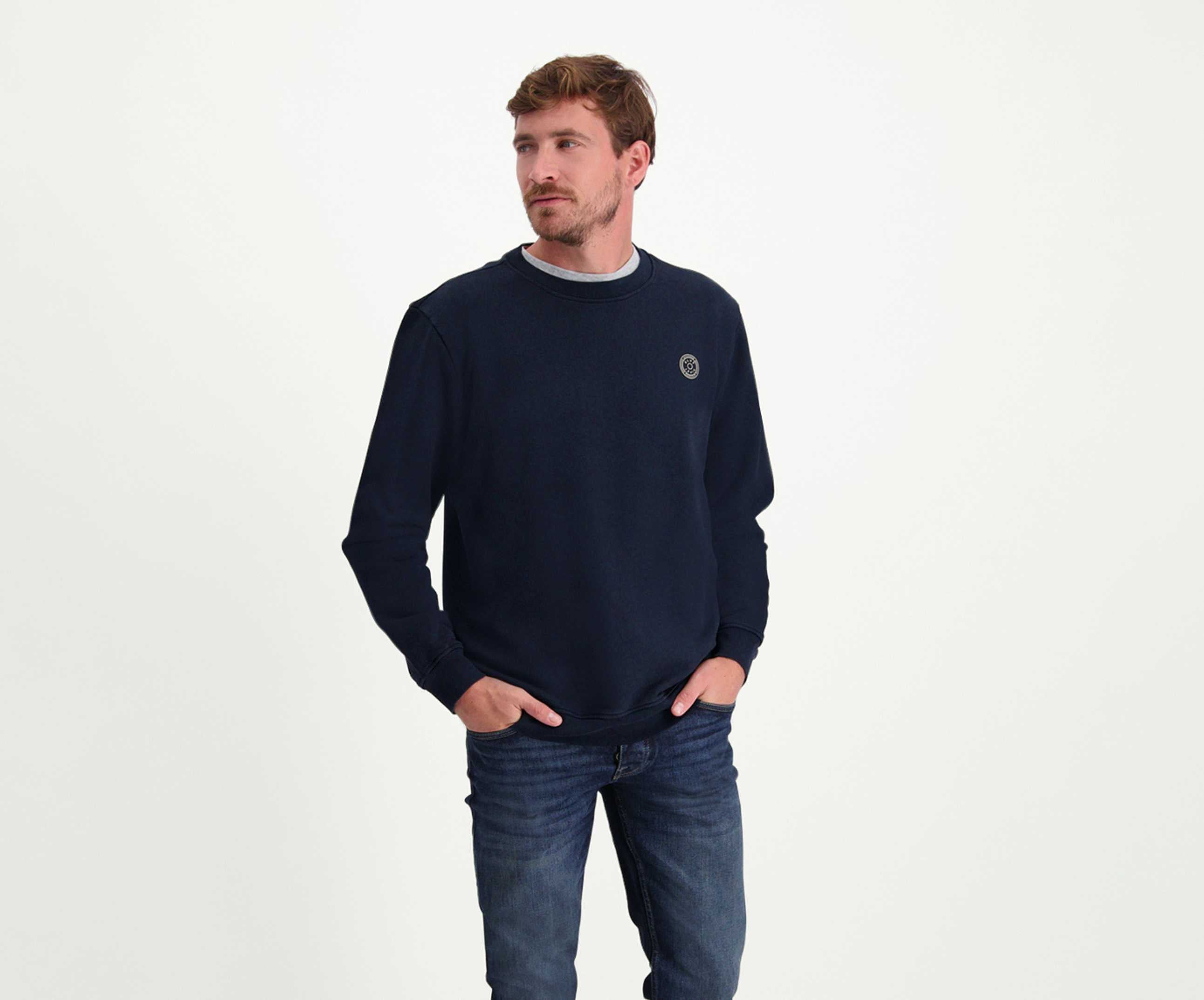 Een man met kort haar draagt The N01 Sweat Navy van Blue Rivet Jeans, gemaakt van biologisch katoen, naast een blauwe spijkerbroek. Hij staat met zijn handen in zijn zakken en kijkt zijwaarts tegen een effen, lichtgekleurde achtergrond die de normale pasvorm van de trui accentueert.