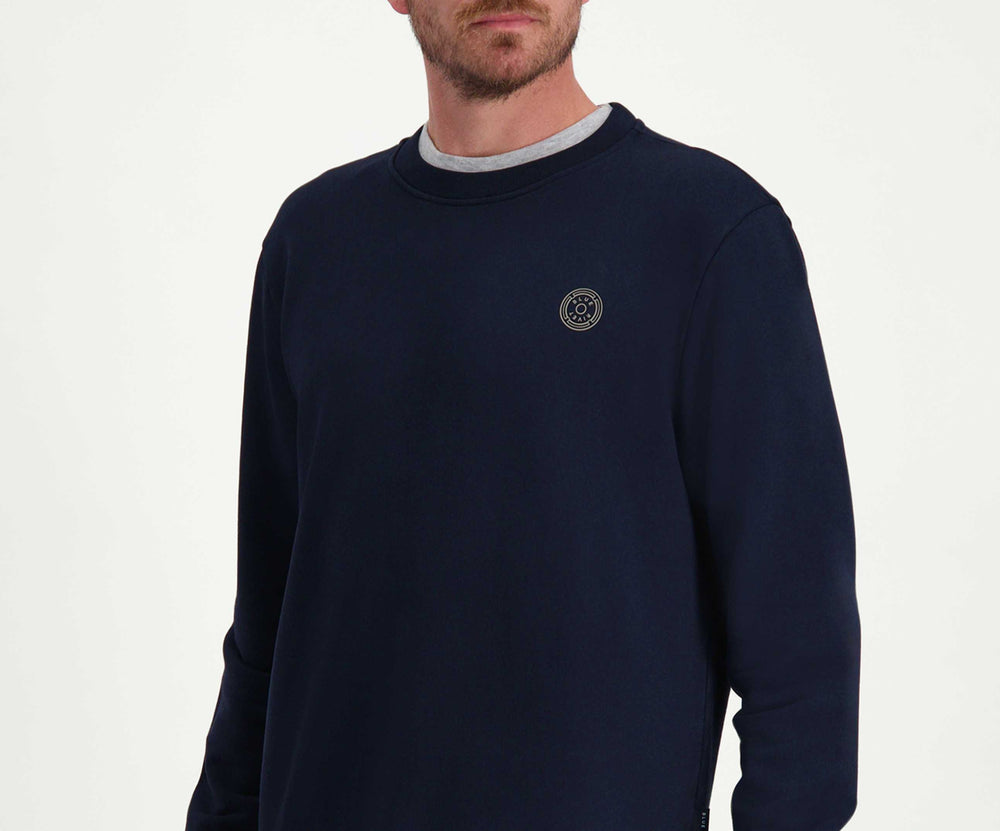 Een persoon met kort bruin haar en een baard draagt *The N01 Sweat Navy* van Blue Rivet Jeans, gemaakt van premium biologisch katoen in een normale pasvorm, gecombineerd met jeans. De persoon kijkt opzij met één hand in een zak, staande tegen een effen witte achtergrond.