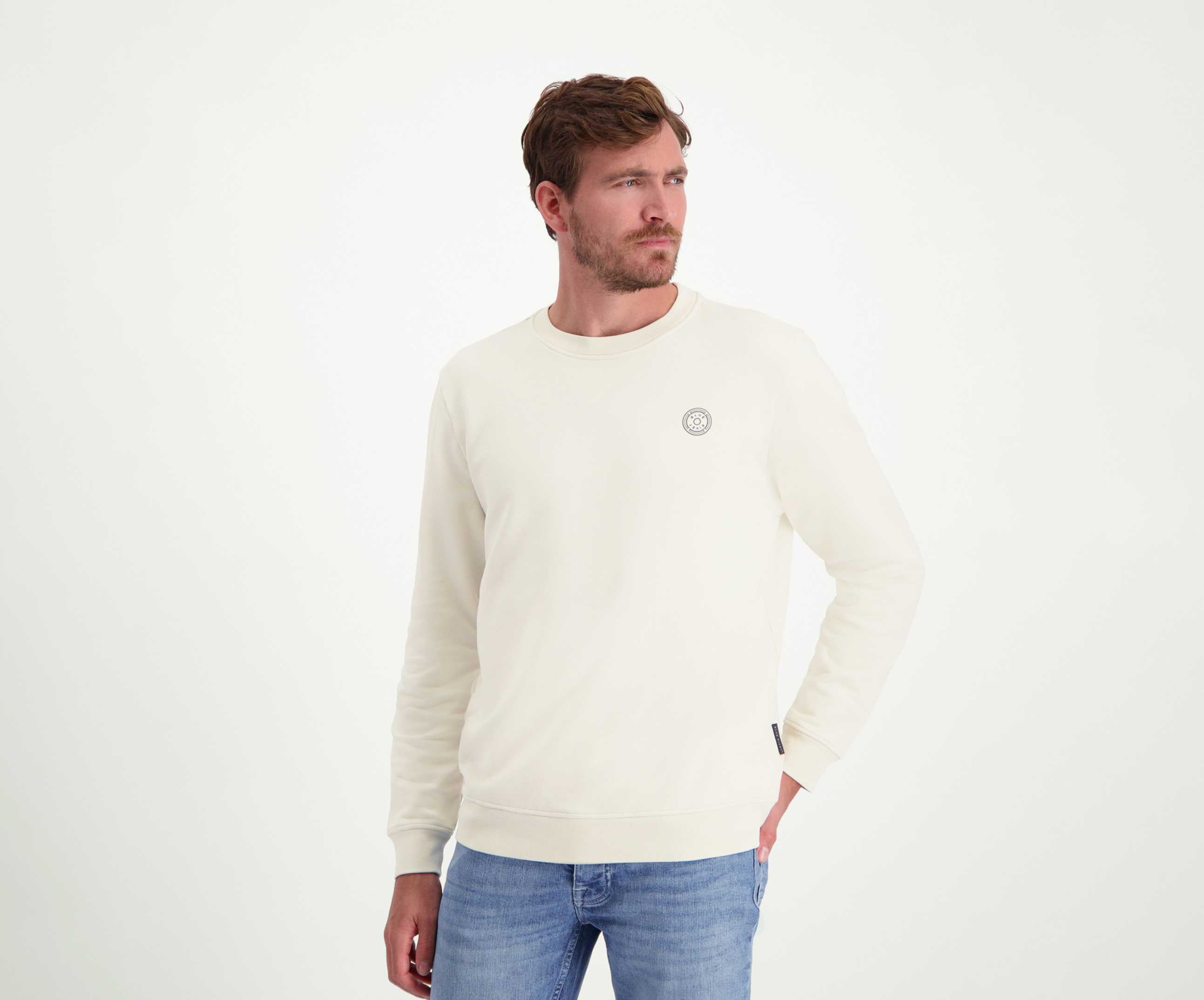 Een man met kort haar en een baard kijkt opzij en draagt "The N01 Sweat Off White" van Blue Rivet Jeans. De trui heeft een klein rond logo op de borst en is gecombineerd met lichtblauwe jeans. De normale pasvorm benadrukt zijn casual stijl tegen de effen witte achtergrond.