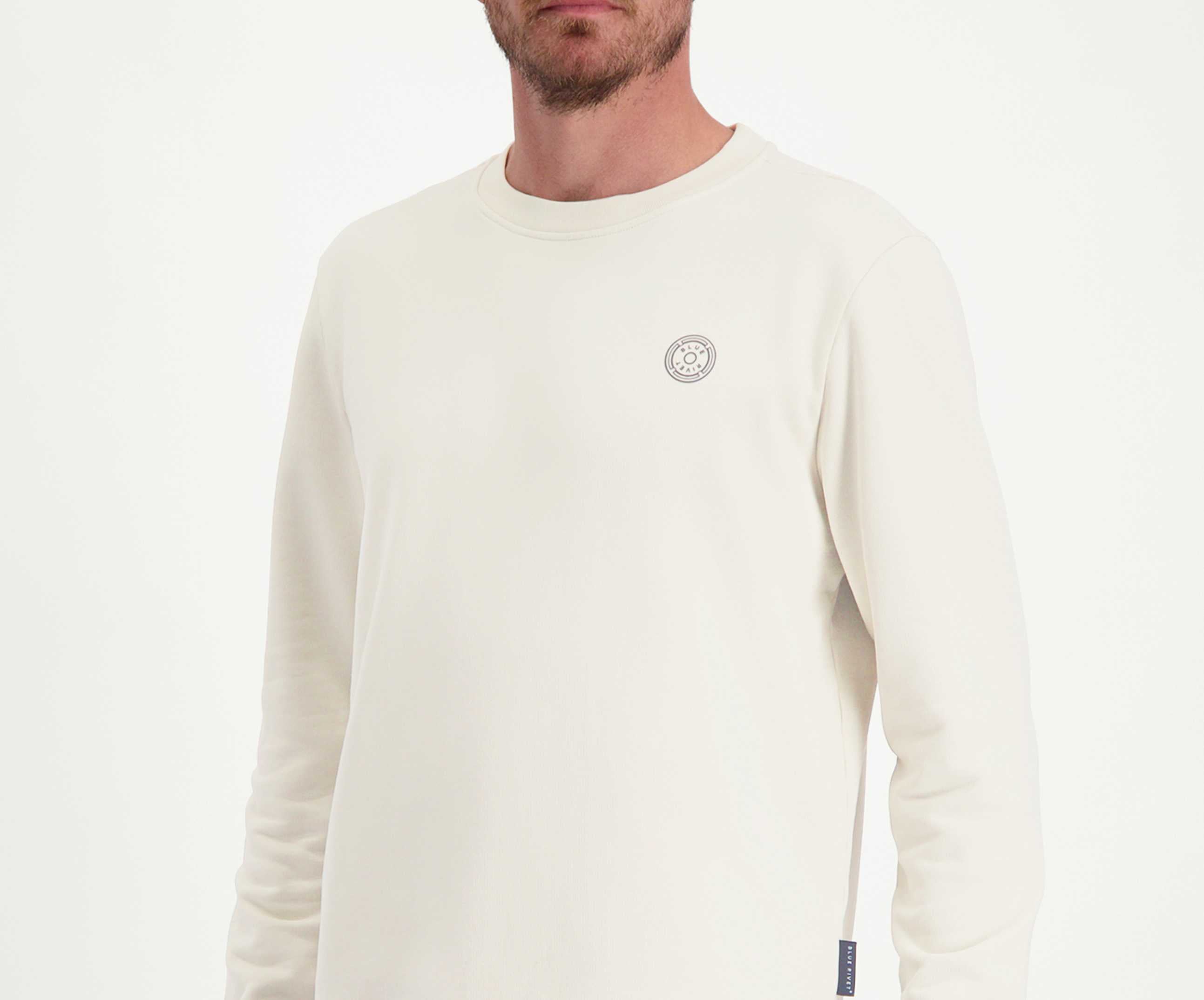 Een man draagt *The N01 Sweat Off White* van Blue Rivet Jeans, een premium trui met lange mouwen gemaakt van biologisch katoen, versierd met een klein rond embleem op de borst, tegen een effen achtergrond.