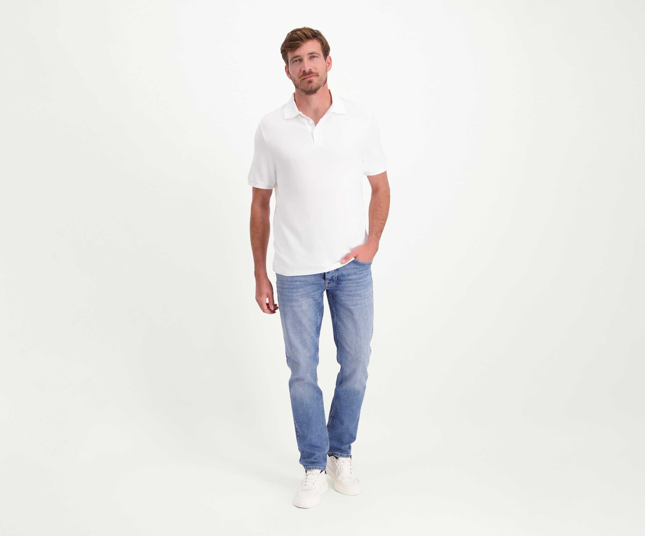 Een man in een wit poloshirt en The N01 Slim Stone Bleached jeans van Blue Rivet Jeans staat tegen een effen lichte achtergrond. Met één hand in zijn zak en premium witte sneakers kijkt hij nonchalant naar de camera.