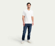 Een man draagt een wit poloshirt, Blue Rivet Jeans' The N01 Straight Dark Indigo Used en witte sneakers tegen een effen, lichte achtergrond.