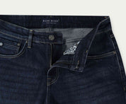 Een paar The N01 Straight Dark Indigo Used jeans met een knoop en een gedeeltelijk open rits. De tailleband heeft een Blue Rivet Jeans-label. Deze denim jeans zijn plat gelegd op een neutrale achtergrond, wat de klassieke stijl benadrukt.