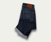 Een paar The N01 Straight Dark Indigo Used jeans van Blue Rivet Jeans, netjes opgevouwen met een zichtbare bruine leren patch op de tailleband. De stretchstof verbetert het comfort en ze worden weergegeven op een lichte achtergrond, waarbij de achterzak en de omgeslagen pijpen worden getoond.