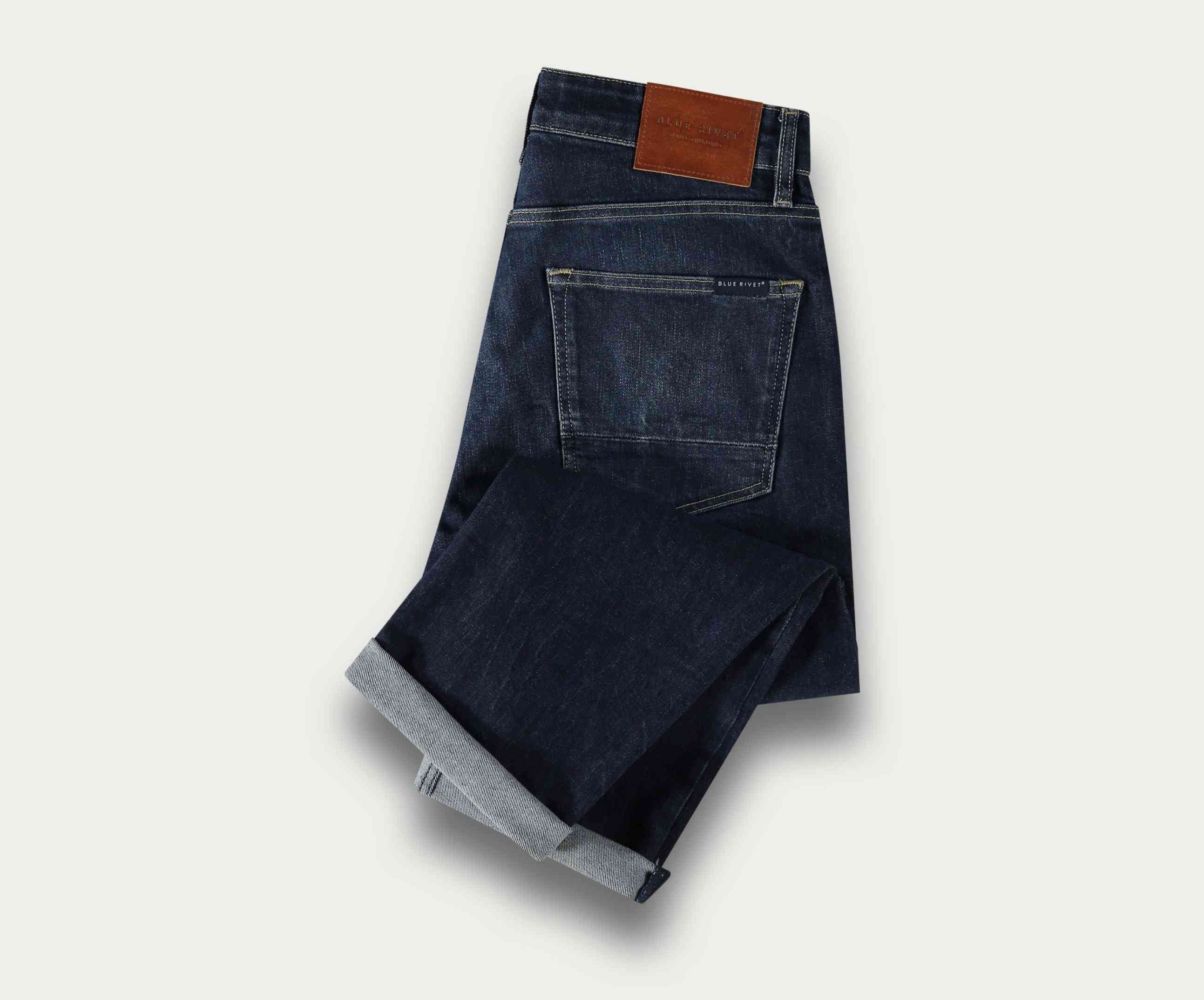 Een paar The N01 Straight Dark Indigo Used jeans van Blue Rivet Jeans, netjes opgevouwen met een zichtbare bruine leren patch op de tailleband. De stretchstof verbetert het comfort en ze worden weergegeven op een lichte achtergrond, waarbij de achterzak en de omgeslagen pijpen worden getoond.