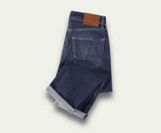 Een netjes opgevouwen paar The N01 Straight Indigo Used jeans van Blue Rivet Jeans, met een bruine leren patch op de tailleband en een zichtbare achterzak, geplaatst op een lichte achtergrond.