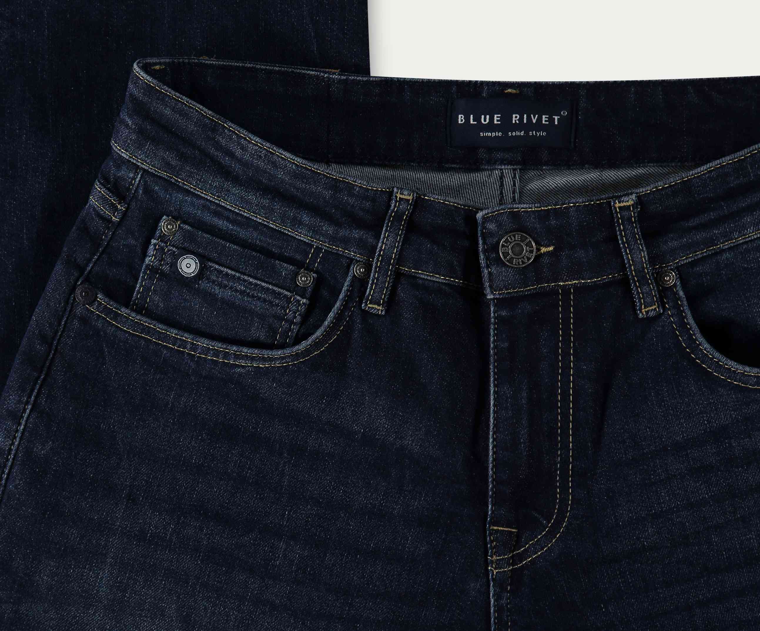 Close-up van donker indigo stretch denim jeans uit de N01 Straight Dark Indigo Used collectie, plat gelegd om de voorkant van de tailleband en zakken te tonen. Het binnenlabel vermeldt Blue Rivet Jeans, met merkdetails. Deze jeans hebben een klassiek design met zichtbare stiksels, wat zorgt voor een rechte pasvorm die comfort en stijl combineert.