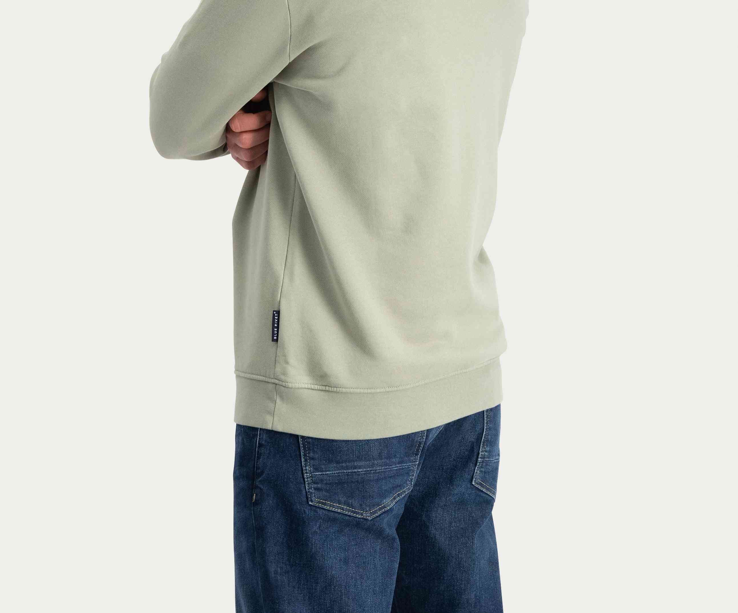 Een persoon die The N01 Sweat Seagrass draagt, gemaakt van biologisch katoen, staat met zijn armen gekruist en kijkt de andere kant op. Hij heeft de trui gecombineerd met een regular fit donkerblauwe jeans van Blue Rivet Jeans tegen een effen witte achtergrond.