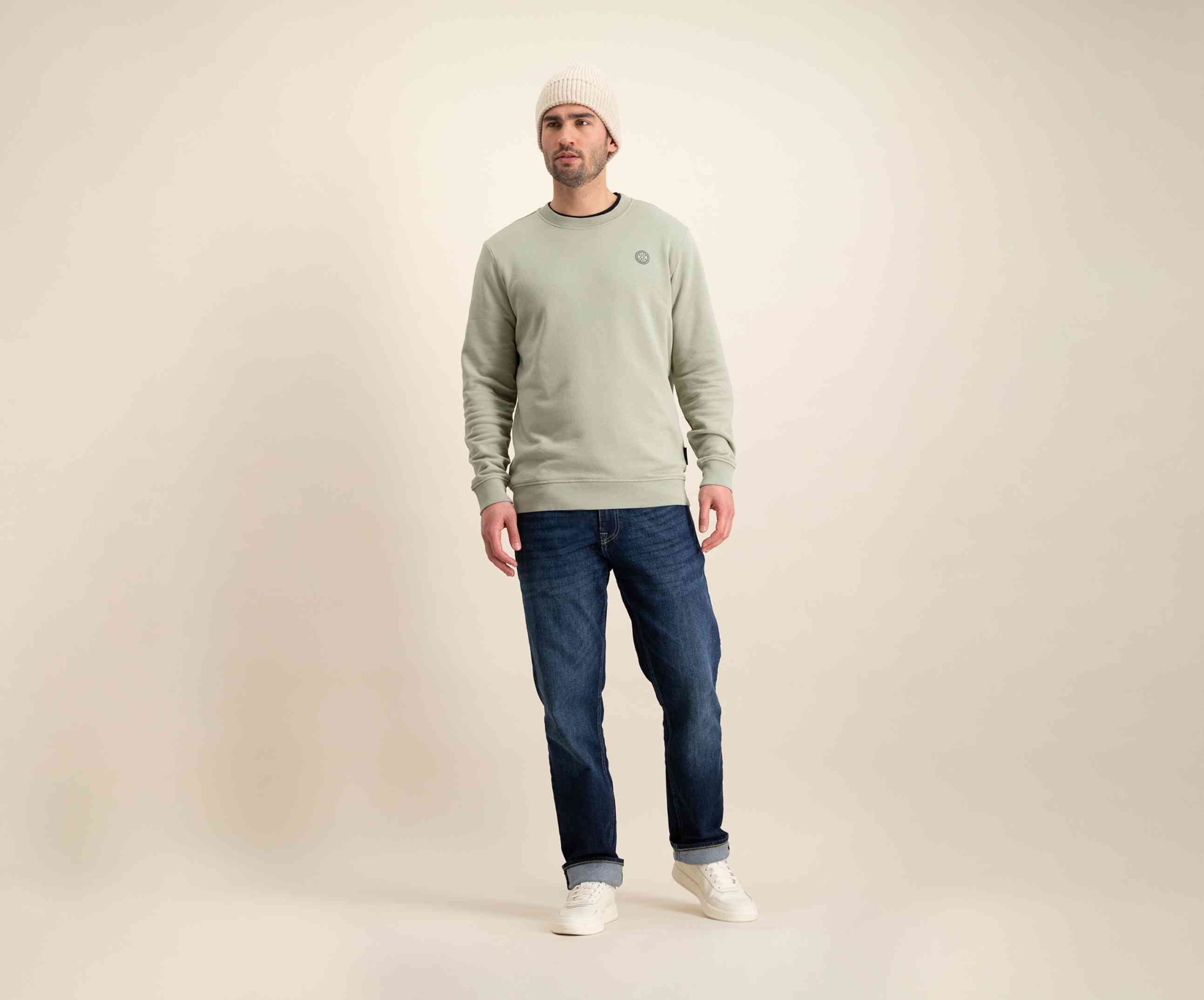 Een man met een beige muts, The N01 Sweat Seagrass van Blue Rivet Jeans, en blauwe jeans staat tegen een effen, lichtgekleurde achtergrond. Hij draagt witte sneakers en kijkt iets opzij, waarmee hij zijn moeiteloos coole stijl laat zien in deze Premium Sweater Heren-look.