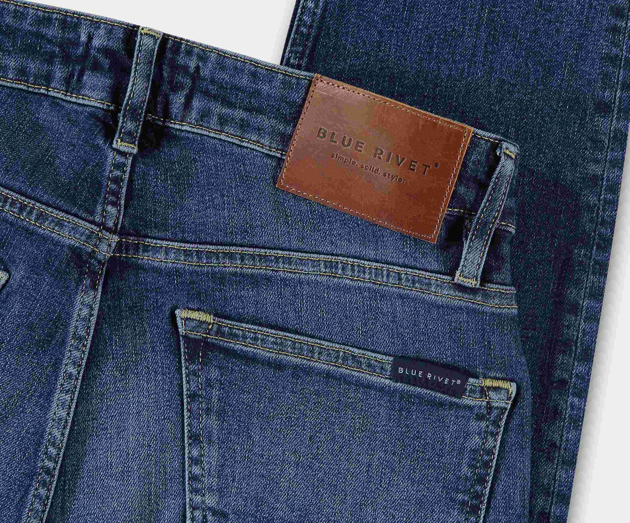 Een donkerblauwe premium denim jeans, The N01 Slim Indigo Used by Blue Rivet Jeans, met een leren patch op de tailleband met hun logo. Deze stretch jeans hebben een zichtbare achterzak en een klein label met bijpassende branding, allemaal plat op een wit oppervlak.