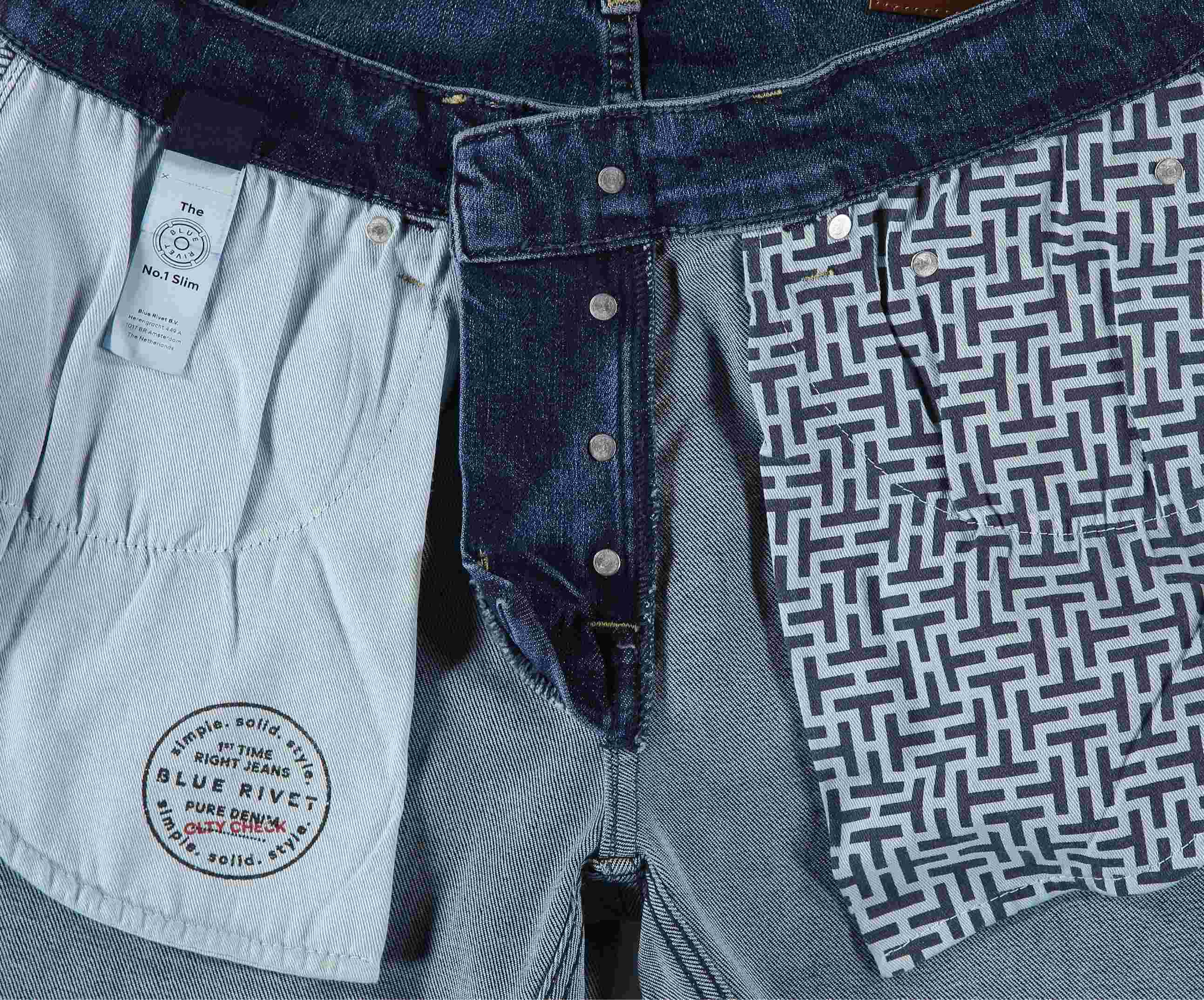 Close-up van de binnenkant van deze premium jeans, met een geometrisch zwart-wit patroonzakje en een effen zakje met een rond label met de tekst The N01 Slim Indigo Used, Blue Rivet Jeans, Pure Denim. De spijkerbroekstof is lichtblauw denim.