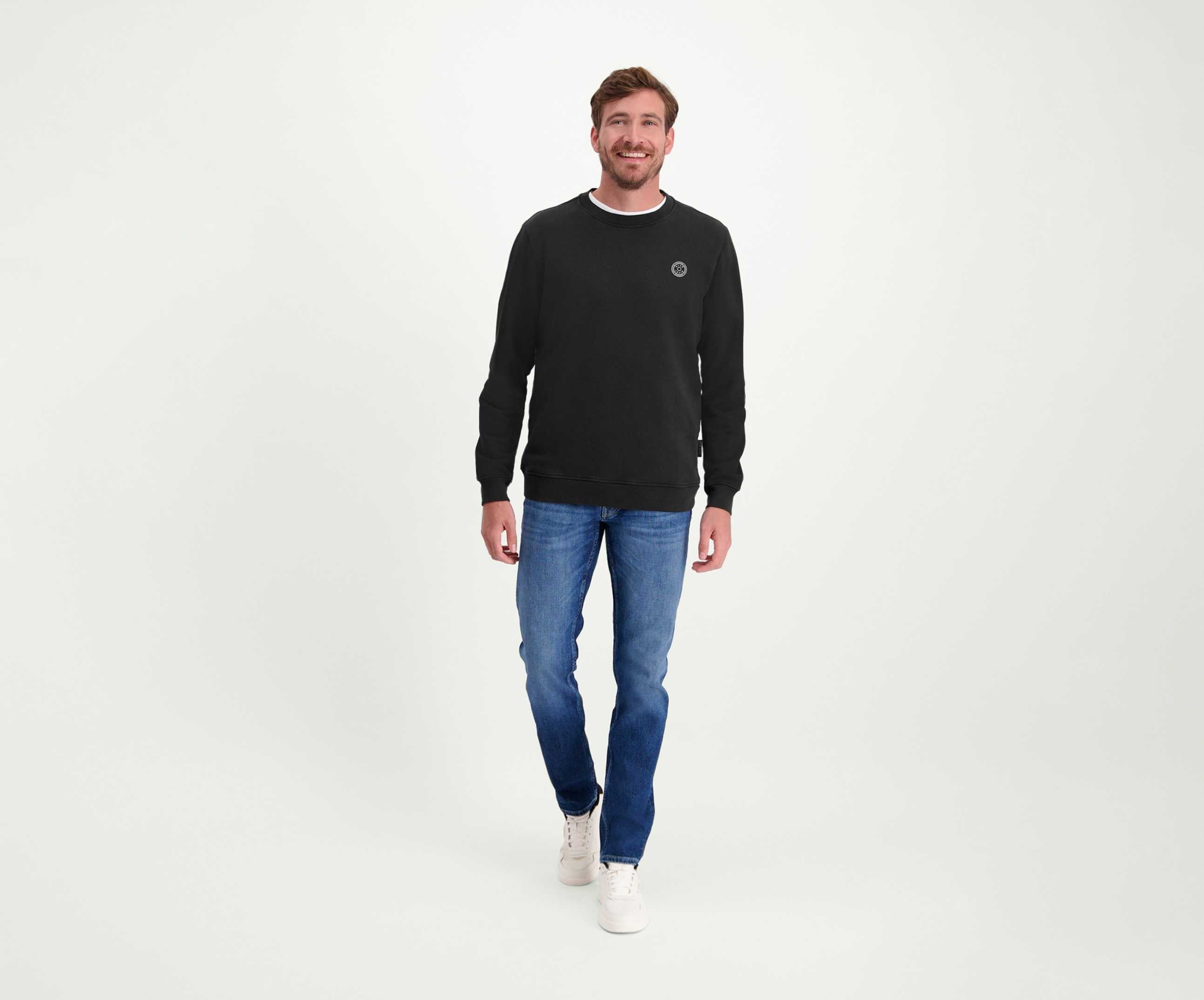 Een man staat in een ontspannen houding, draagt een zwarte trui, The N01 Slim Indigo Used jeans van Blue Rivet Jeans en witte sneakers. De achtergrond is effen wit. Hij heeft een lichte glimlach en zijn handen zijn nonchalant langs zijn lichaam.