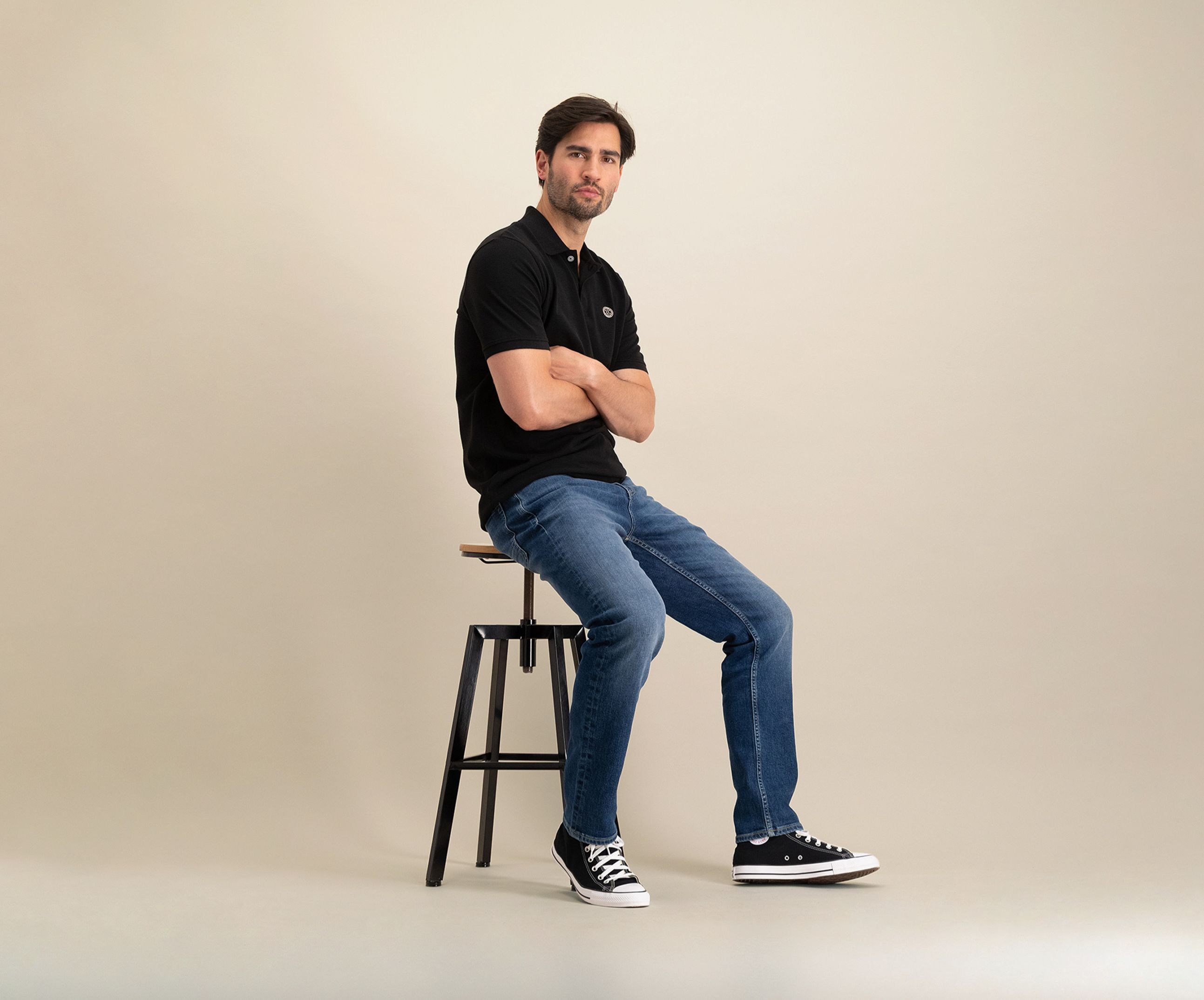 Een man zit op een zwarte kruk tegen een effen beige achtergrond, en straalt zomerse chic uit. Hij draagt The N01 Polo Anthracite, een klassieke polo van biologisch katoen, gecombineerd met de blauwe denim van Blue Rivet Jeans en stijlvolle zwarte sneakers. Met zijn armen over elkaar kijkt hij recht in de camera, wat een moeiteloze stijl uitstraalt.