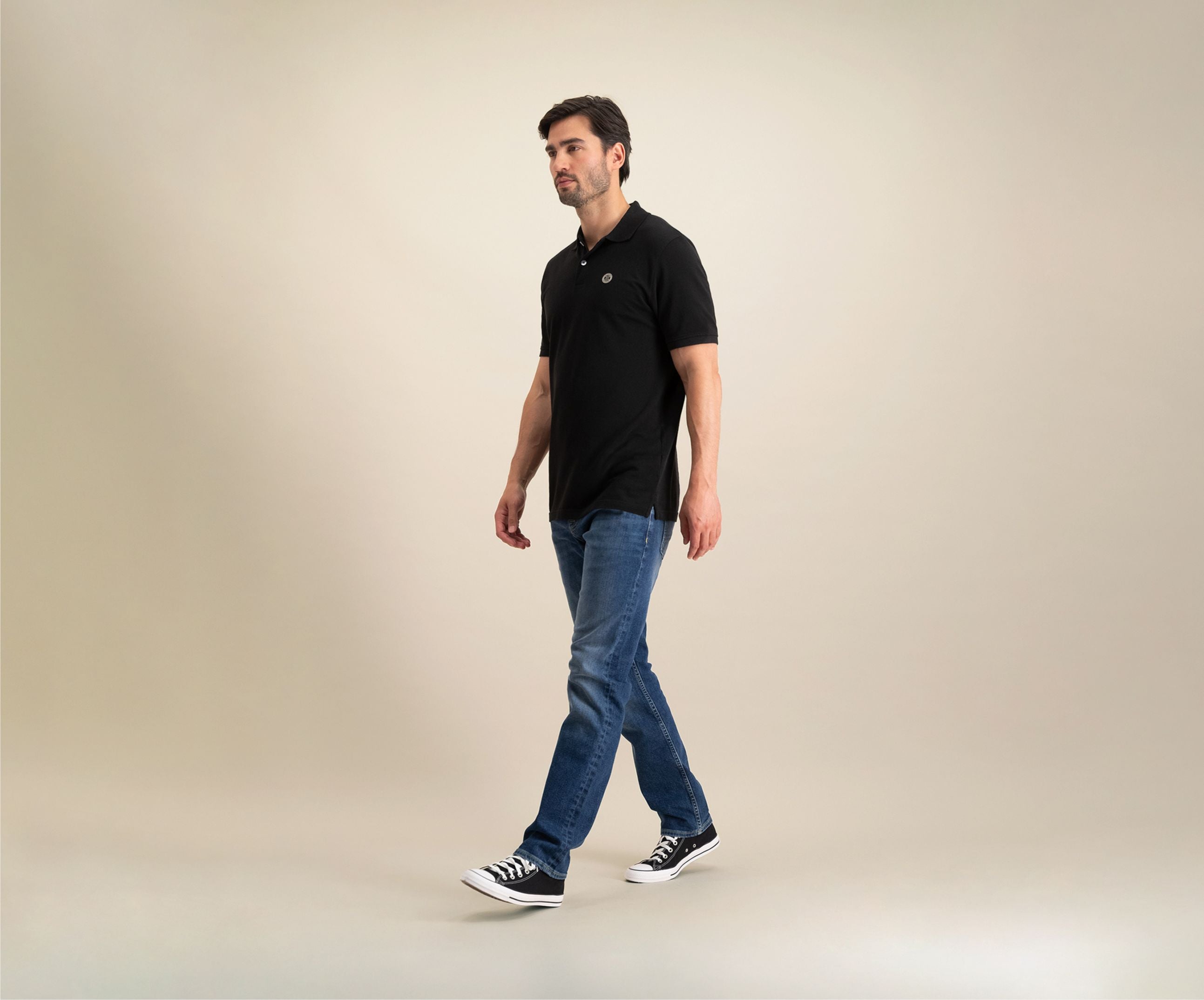 Een man die The N01 Polo Anthracite en Blue Rivet Jeans draagt, loopt zelfverzekerd over een effen, zacht verlichte achtergrond. Met zwarte en witte sneakers straalt hij zomerse chic uit terwijl hij met een ontspannen houding naar links kijkt.