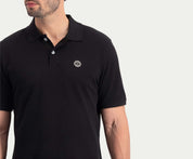 Een man staat tegen een effen witte achtergrond en draagt The N01 Polo Anthracite van Blue Rivet Jeans, gemaakt van biologisch katoen en met een discreet rond logo op de borst. Het is perfect om die chique zomerlook te bereiken.