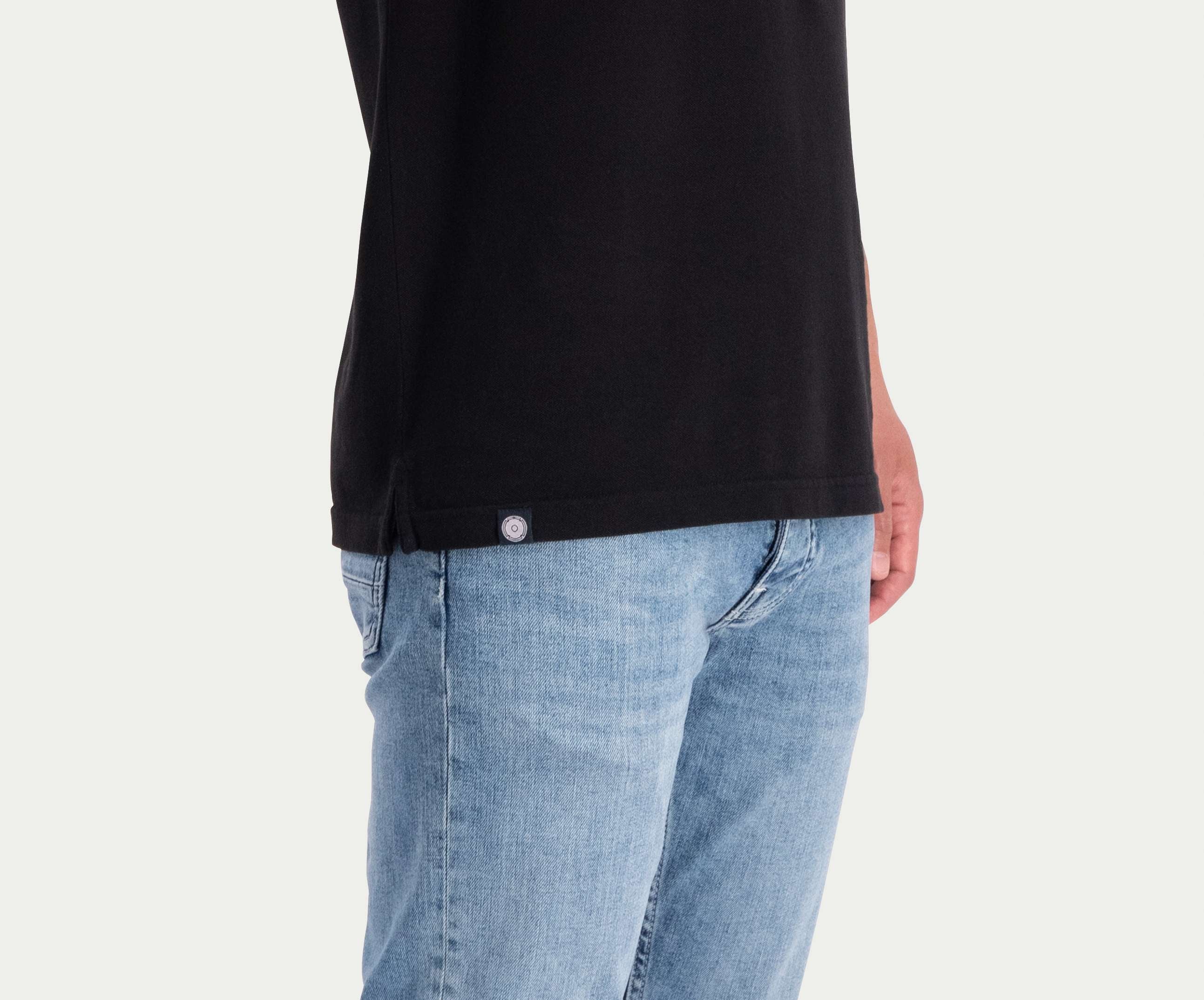 Een persoon die The N01 Polo Anthracite and Blue Rivet Jeans draagt, in een lichtblauwe wassing, staat tegen een effen achtergrond. Alleen het onderste deel van het poloshirt en de jeans met een klein label zichtbaar op het shirt worden getoond, wat een vleugje zomerse chic toevoegt aan hun ingetogen look.