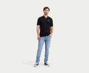 Een persoon staat tegen een effen, lichte achtergrond en draagt The N01 Polo Anthracite, lichtblauwe jeans van Blue Rivet Jeans en zwart-witte sneakers. Eén hand zit in hun zak en ze hebben een ontspannen uitdrukking die zomerse chic uitstraalt.