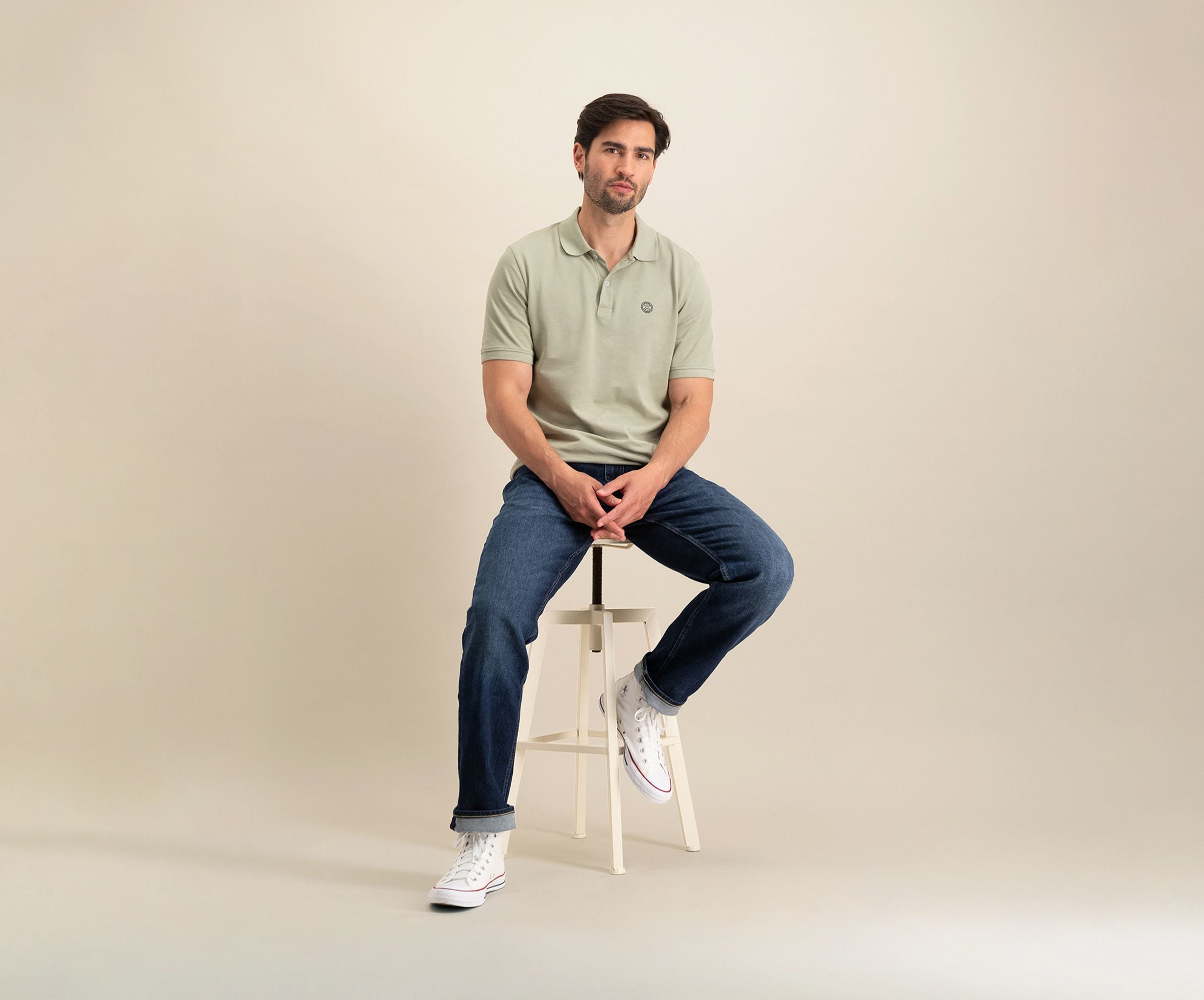 Een man met kort haar en een baard zit op een kruk tegen een neutrale achtergrond. Hij draagt "The N01 Polo Seagrass" en donkerblauwe jeans van Blue Rivet Jeans, gecombineerd met witte high-top sneakers, kijkt recht vooruit met een ontspannen houding.