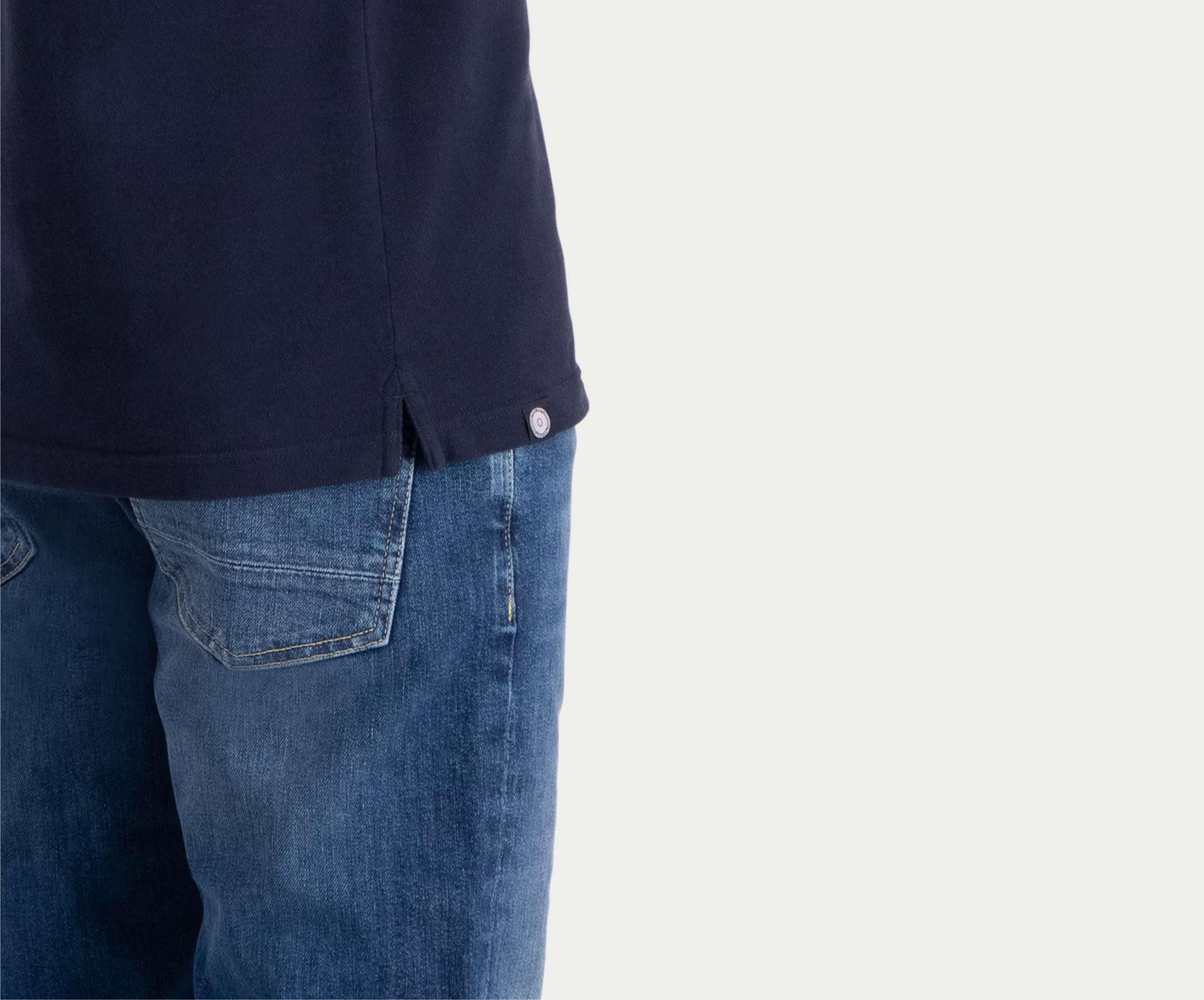 Een persoon die gedeeltelijk zichtbaar is, draagt The N01 Polo Navy samen met Blue Rivet Jeans. Het shirt heeft een klein knoopdetail bij een zijsplit en de achtergrond is effen gebroken wit.