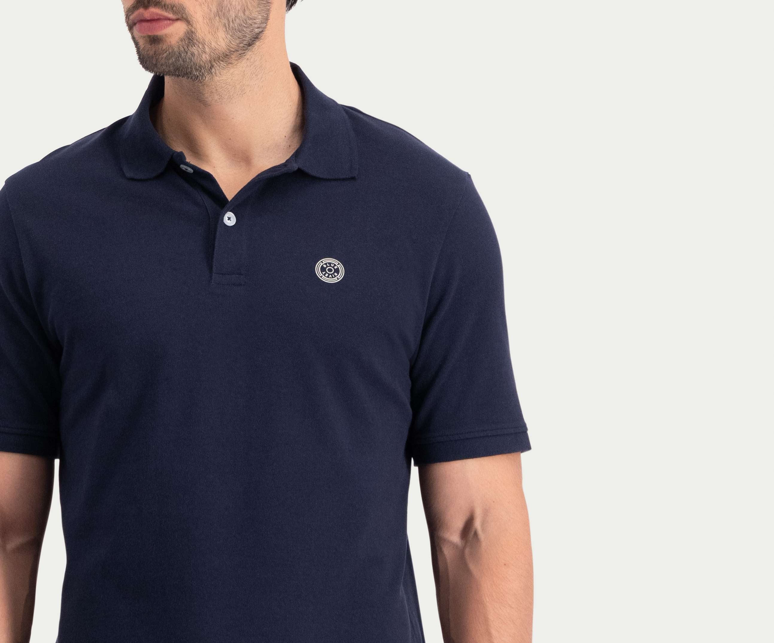 Een persoon die The N01 Polo Navy van Blue Rivet Jeans draagt, met korte mouwen en een klein rond logo op de borst, staat tegen een effen witte achtergrond. Het shirt heeft een klassieke kraag en het gezicht van de persoon is gedeeltelijk buiten beeld.