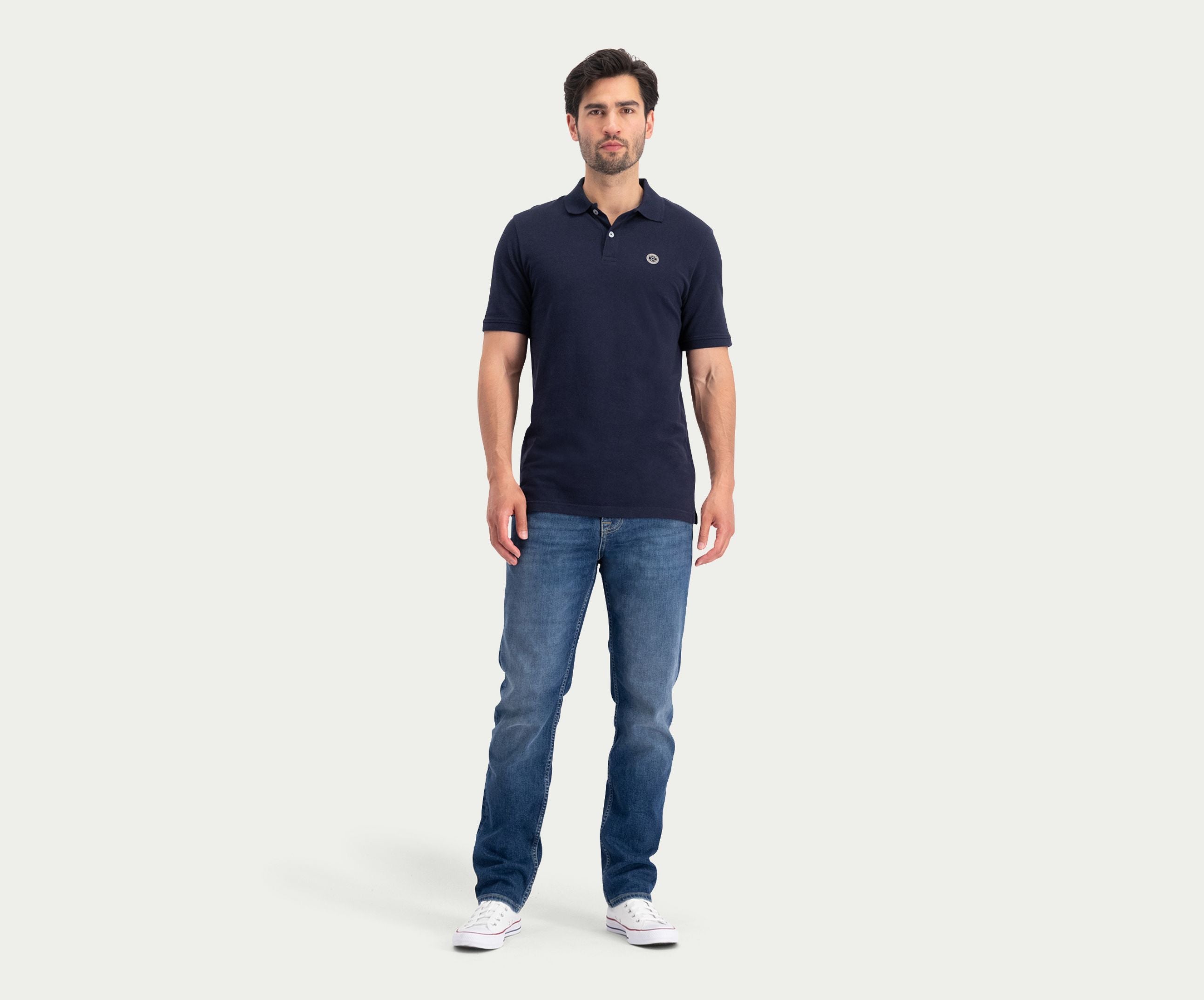 Een man staat voorovergebogen tegen een effen achtergrond, gekleed in de N01 Polo Navy, Blue Rivet Jeans en witte sneakers.