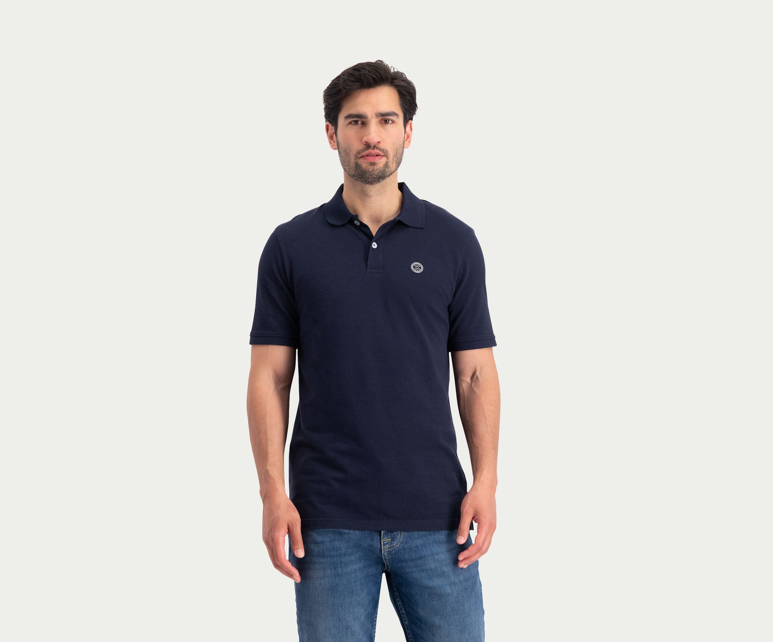 Een man met donker haar en een baard draagt de N01 Polo Navy, gecombineerd met Blue Rivet Jeans, en staat tegen een lichte, effen achtergrond.