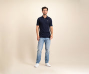 Tegen een effen beige achtergrond is een man te zien in The N01 Polo Navy en lichtblauwe Blue Rivet Jeans, aangevuld met witte sneakers. Hij heeft kort donker haar en een neutrale uitdrukking.