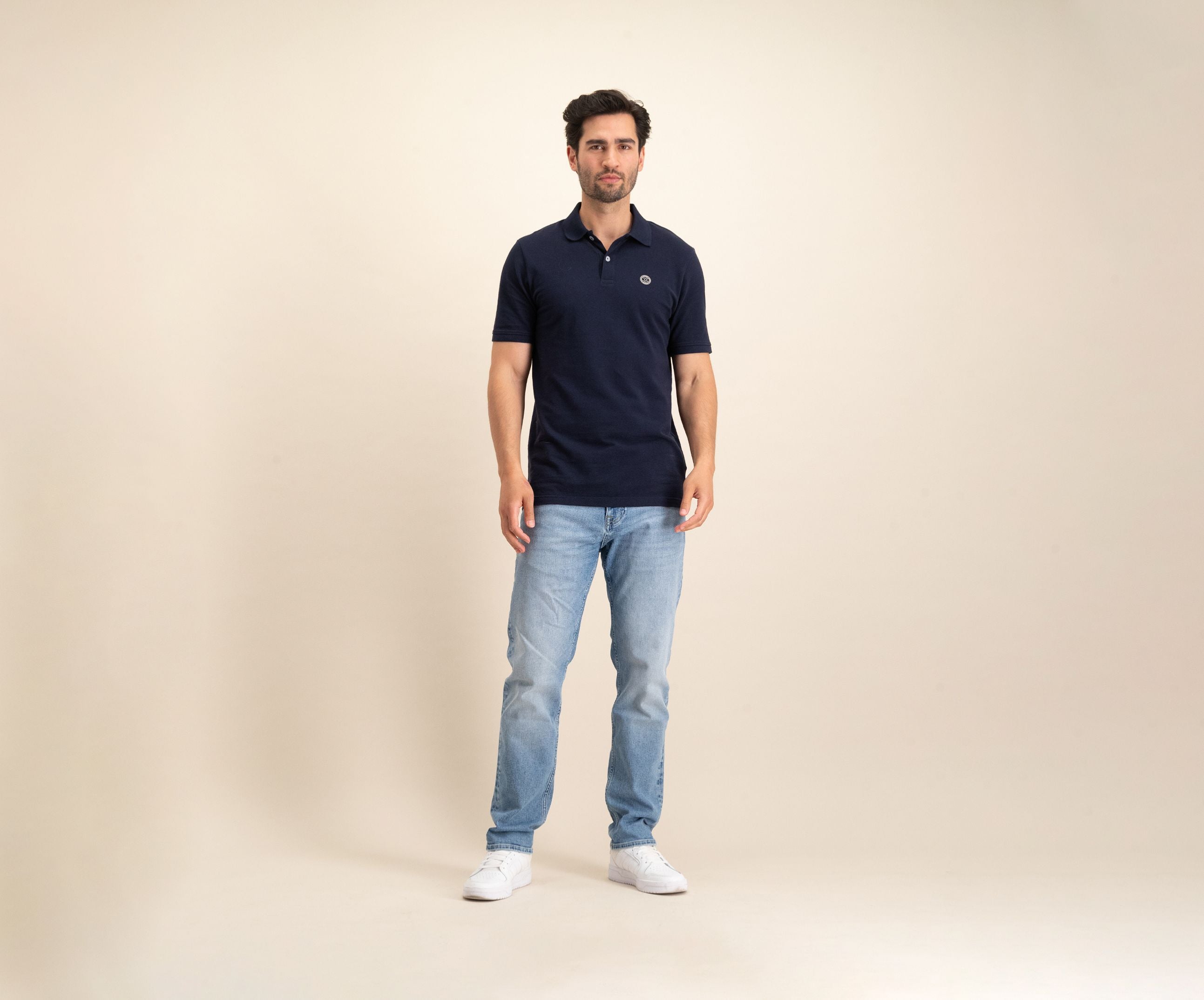 Tegen een effen beige achtergrond is een man te zien in The N01 Polo Navy en lichtblauwe Blue Rivet Jeans, aangevuld met witte sneakers. Hij heeft kort donker haar en een neutrale uitdrukking.