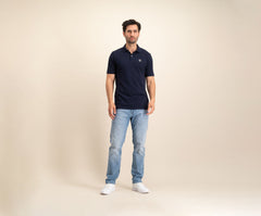 Tegen een effen beige achtergrond is een man te zien in The N01 Polo Navy en lichtblauwe Blue Rivet Jeans, aangevuld met witte sneakers. Hij heeft kort donker haar en een neutrale uitdrukking.