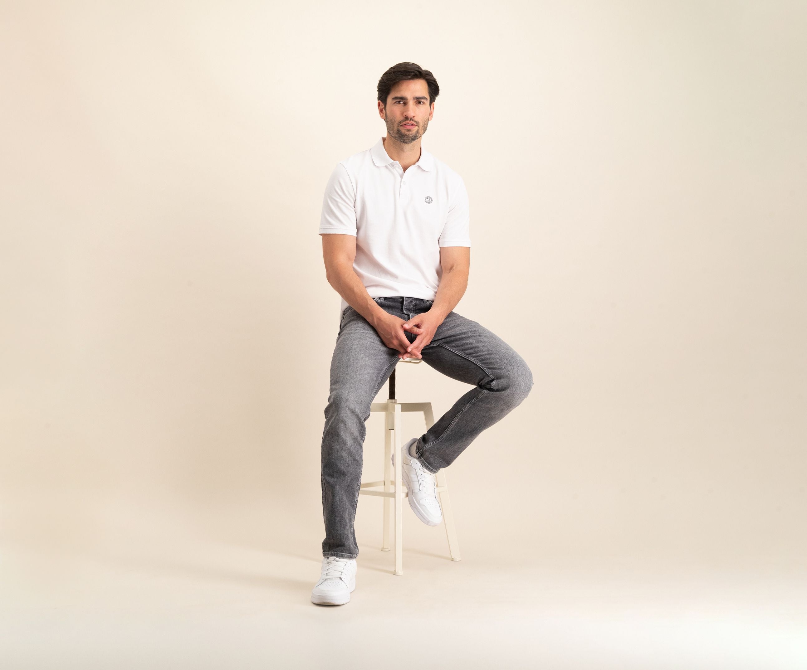 Een man die de N01 Polo White en grijze Blue Rivet Jeans draagt, samen met witte sneakers, zit op een kruk tegen een lichtbeige achtergrond. Hij lijkt ontspannen met één voet op de vloer en zijn handen rustend op zijn schoot.