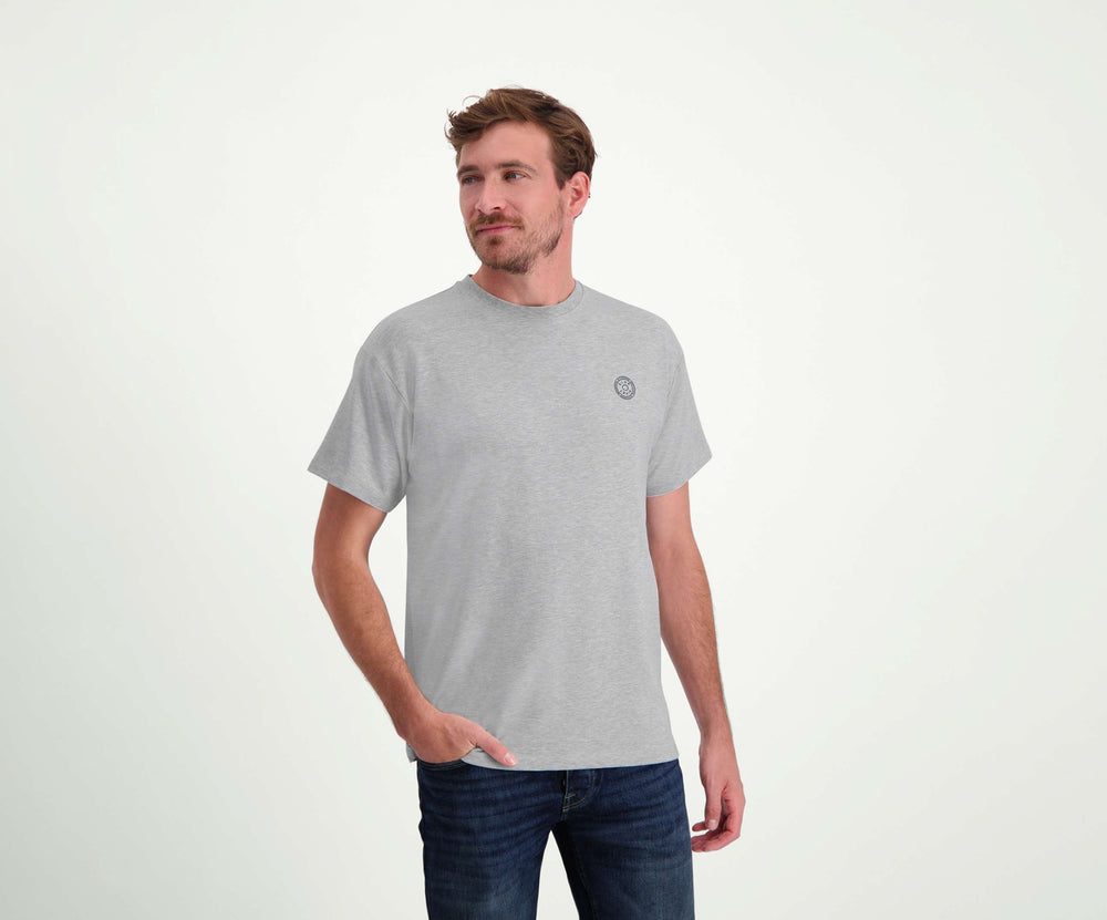 Een man met kort haar staat in de regular fit "The N01 Tee Grey Melee" en Blue Rivet Jeans. Hij heeft een baard en zijn rechterhand zit nonchalant in zijn zak tegen een effen witte muurachtergrond.