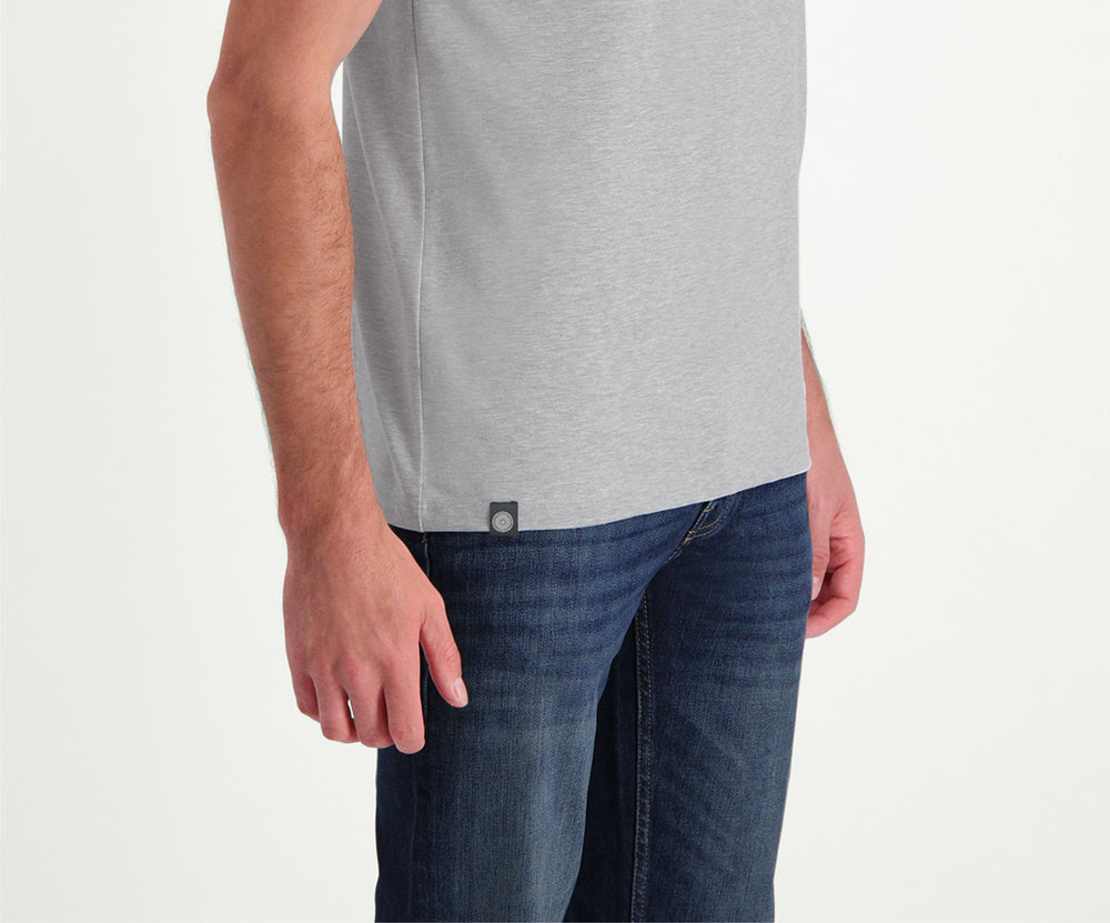 Een man met kort haar staat in de regular fit "The N01 Tee Grey Melee" en Blue Rivet Jeans. Hij heeft een baard en zijn rechterhand zit nonchalant in zijn zak tegen een effen witte muurachtergrond.
