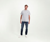 Een man die een The N01 Tee in Grey Melee draagt, staat tegen een effen achtergrond, gecombineerd met Blue Rivet Jeans en witte sneakers. Zijn korte, golvende haar complimenteert zijn ontspannen houding terwijl hij zelfverzekerd in de camera kijkt.