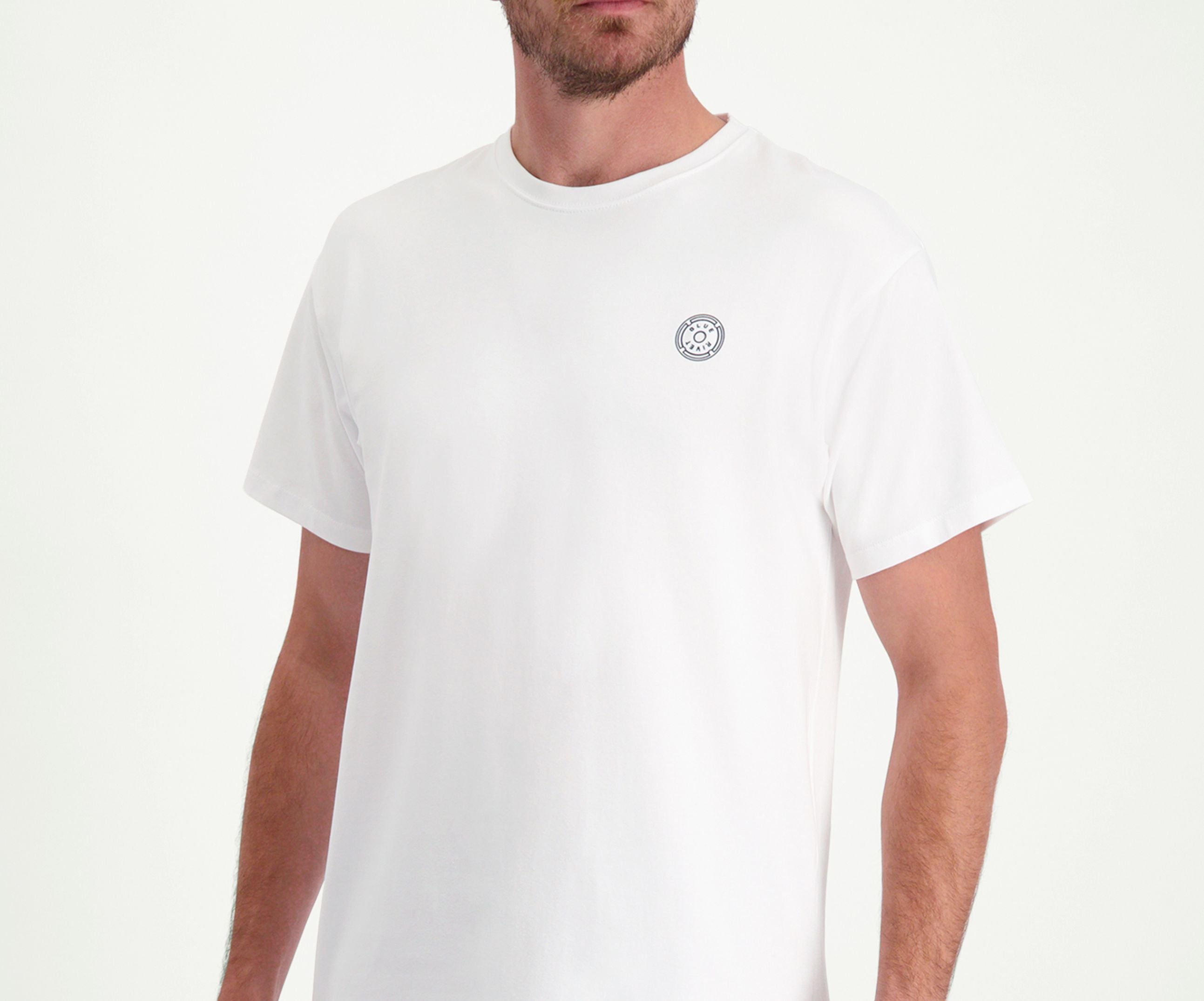 Een persoon die The N01 Tee White van Blue Rivet Jeans draagt, staat tegen een lichte achtergrond. Het T-shirt is regular fit en effen wit, met een klein rond logo op de borst, terwijl het hoofd van de persoon uit het zicht blijft.