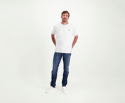 Een man in een regular fit The N01 Tee White and Blue Rivet Jeans staat tegen een effen witte achtergrond. Hij heeft zijn handen in zijn zakken en draagt witte sneakers.