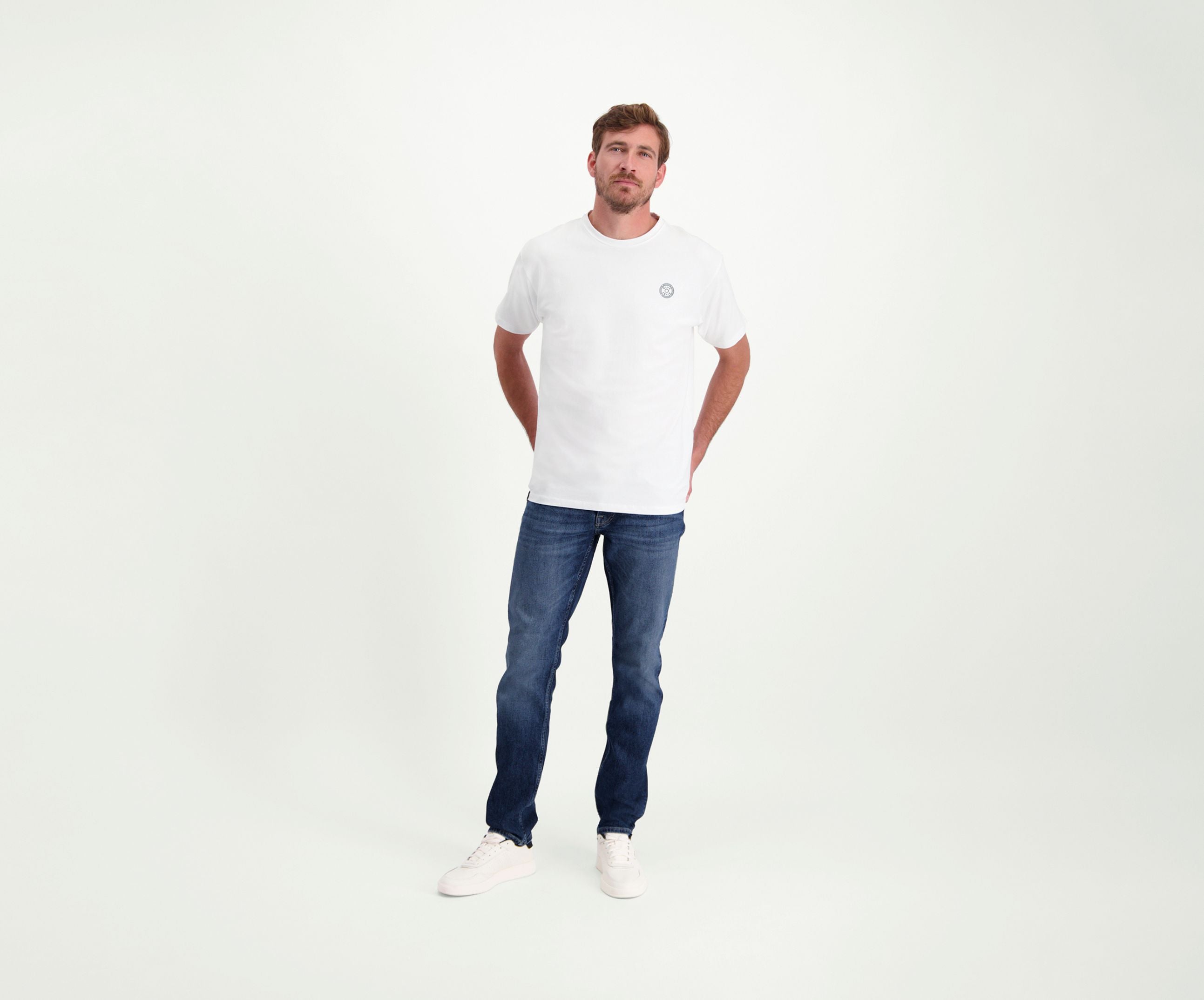 Een man in een regular fit The N01 Tee White and Blue Rivet Jeans staat tegen een effen witte achtergrond. Hij heeft zijn handen in zijn zakken en draagt witte sneakers.