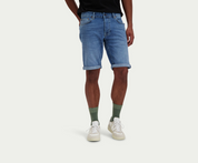 Een persoon die een indigo paar The N01 Short van Blue Rivet Jeans draagt, staat tegen een effen achtergrond. Ze zijn ook gekleed in een zwart shirt, groene sokken en witte sneakers, waarbij de shorts stijlvol zijn opgerold aan de onderkant.