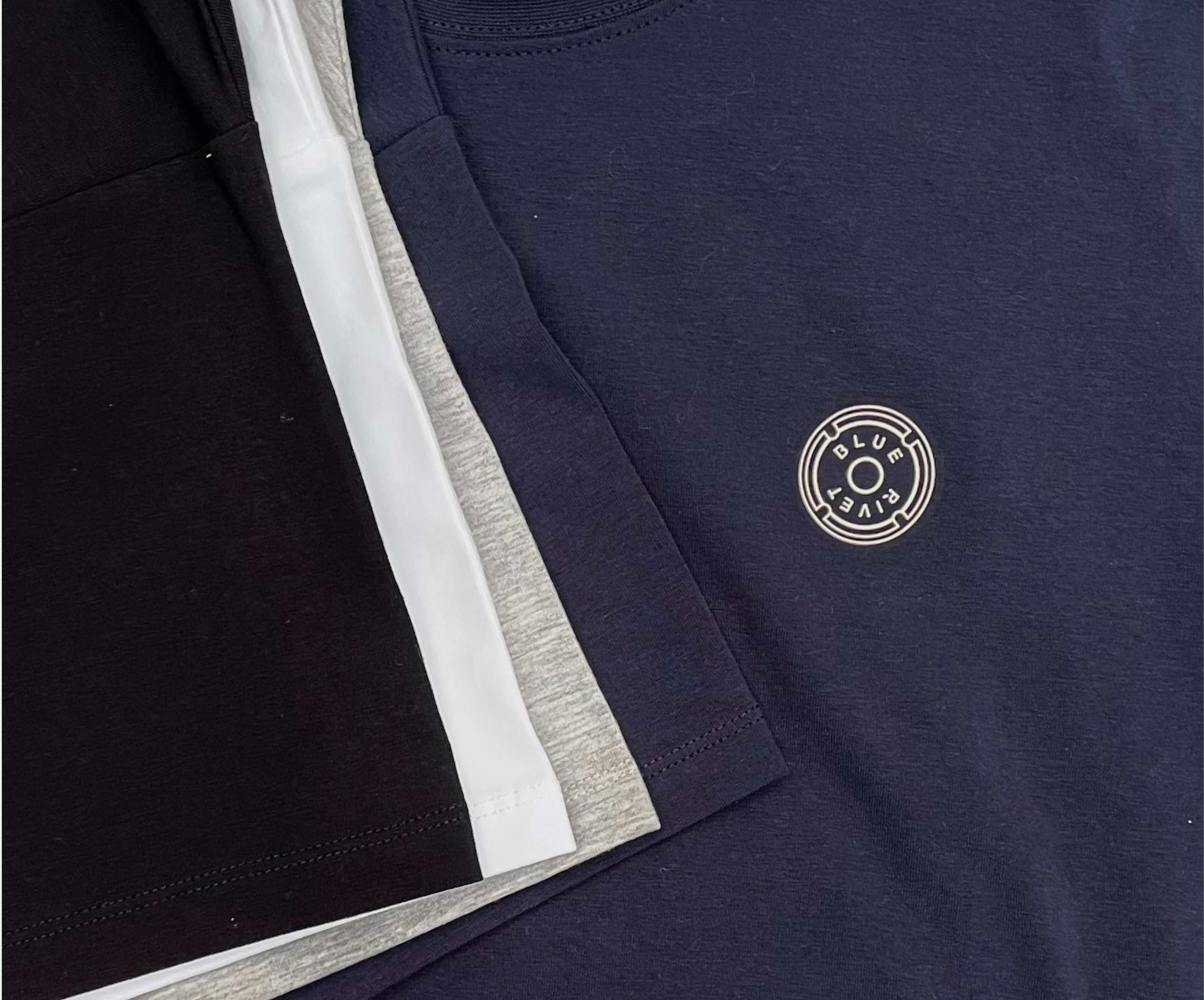 Een collectie Regular Fit gevouwen t-shirts in zwart, wit, grijs en marineblauw. De N01 Tee Navy van Blue Rivet Jeans toont een cirkelvormig embleem met tekst.