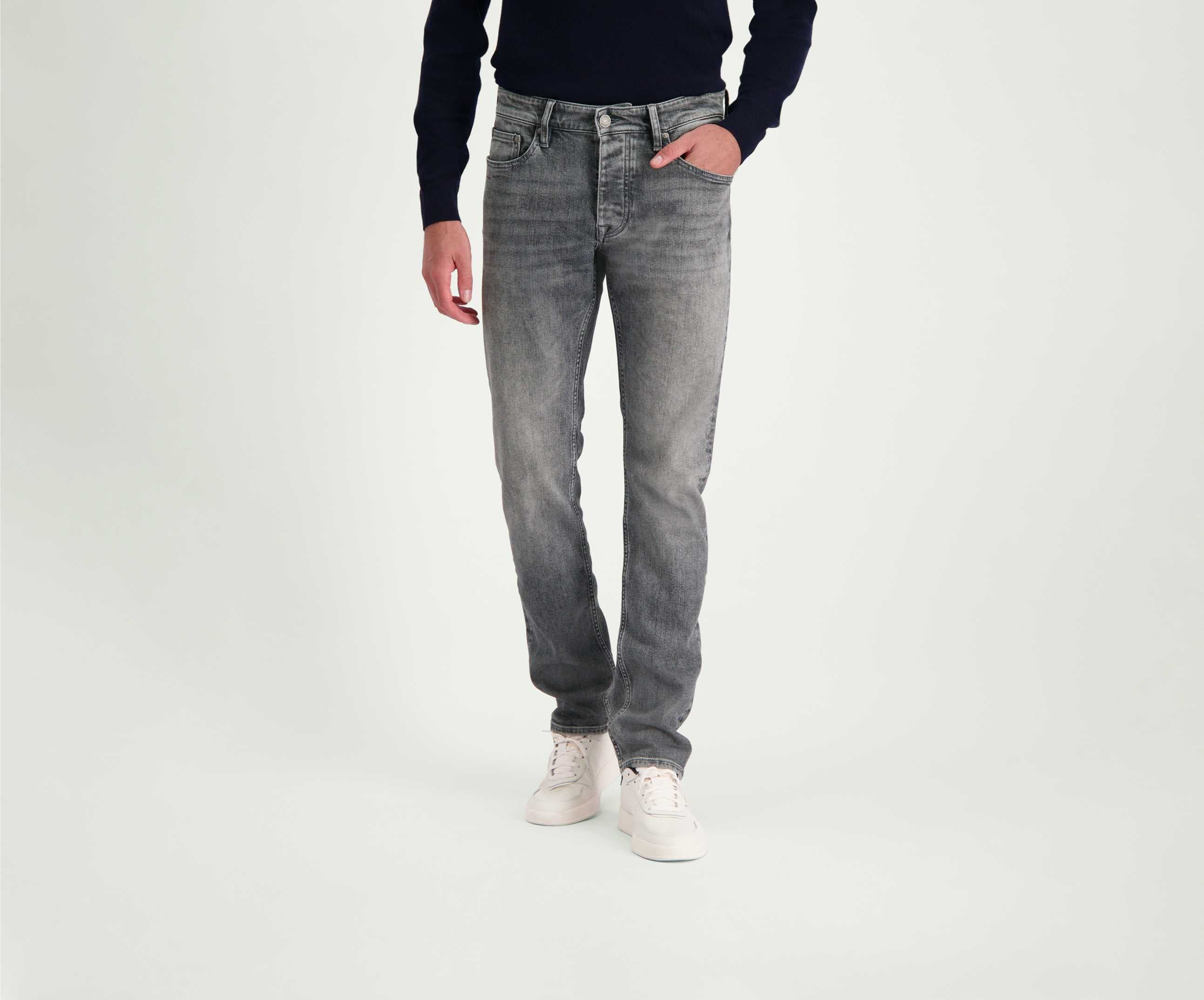 Een persoon staat tegen een neutrale achtergrond en draagt een zwarte trui van topkwaliteit, de N01 Slim Light Grey Used jeans van Blue Rivet Jeans en witte sneakers. Hij heeft één hand in een zak.