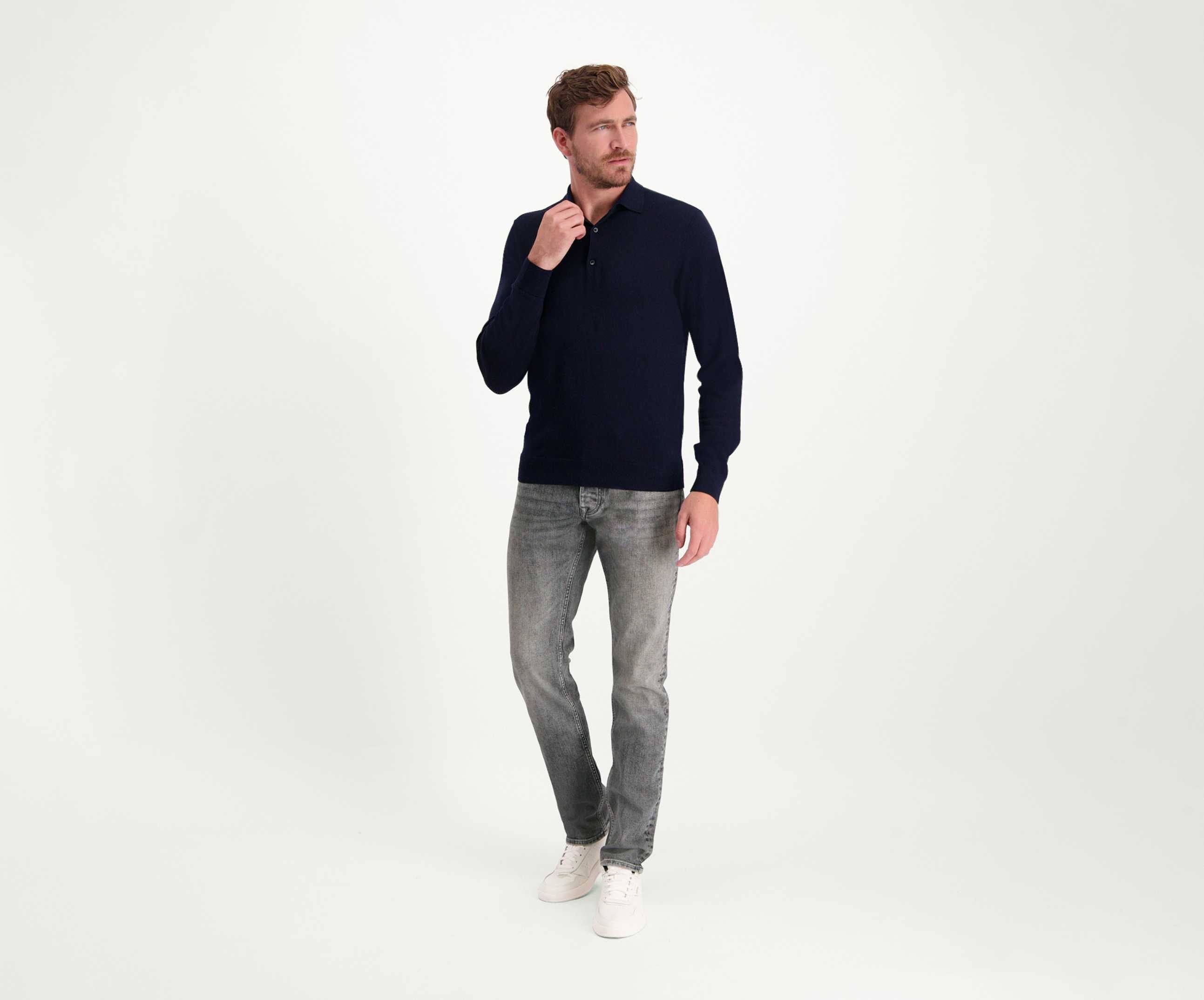 Een man met licht haar staat tegen een effen witte achtergrond, gekleed in een premium donker shirt met lange mouwen, The N01 Slim Light Grey Used jeans van Blue Rivet Jeans en witte sneakers. Hij houdt zijn kraag vast met één hand en kijkt peinzend naar de zijkant.