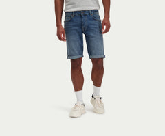 Een persoon draagt een lichtgrijs t-shirt, The N01 Short Dark Indigo Vintage van Blue Rivet Jeans met een opgerolde zoom, witte sokken en witte sneakers. Ze staan tegen een effen witte achtergrond.