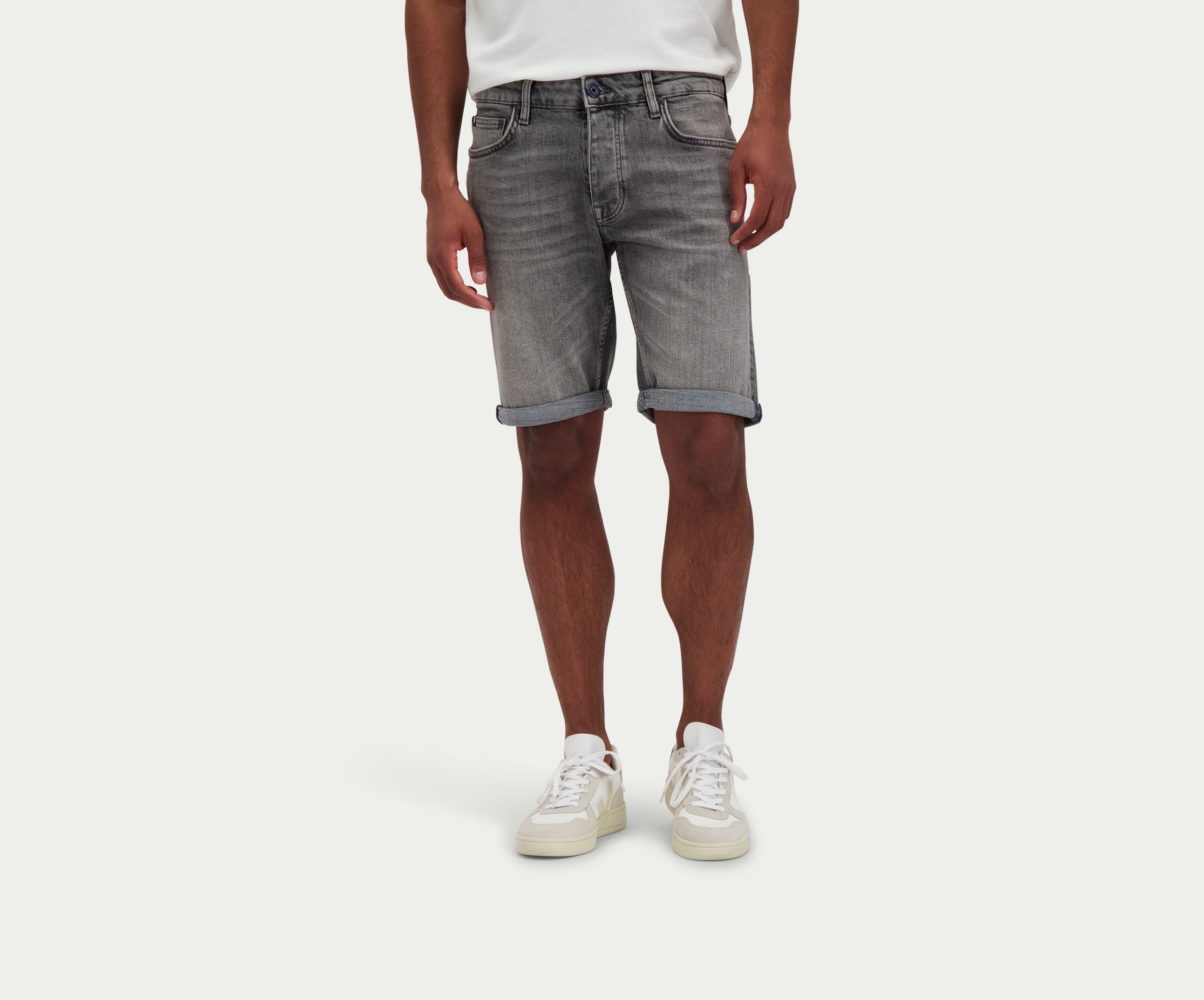Een persoon die een wit t-shirt draagt, The N01 Short Light Grey Used by Blue Rivet Jeans, en witte sneakers staat tegen een effen achtergrond. De shorts zijn opgerold bij de zoom.