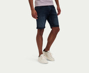 Een persoon die The N01 Short Blue Black van Blue Rivet Jeans draagt en witte sneakers staat tegen een effen witte achtergrond. Alleen de onderkant van het lichaam is zichtbaar.