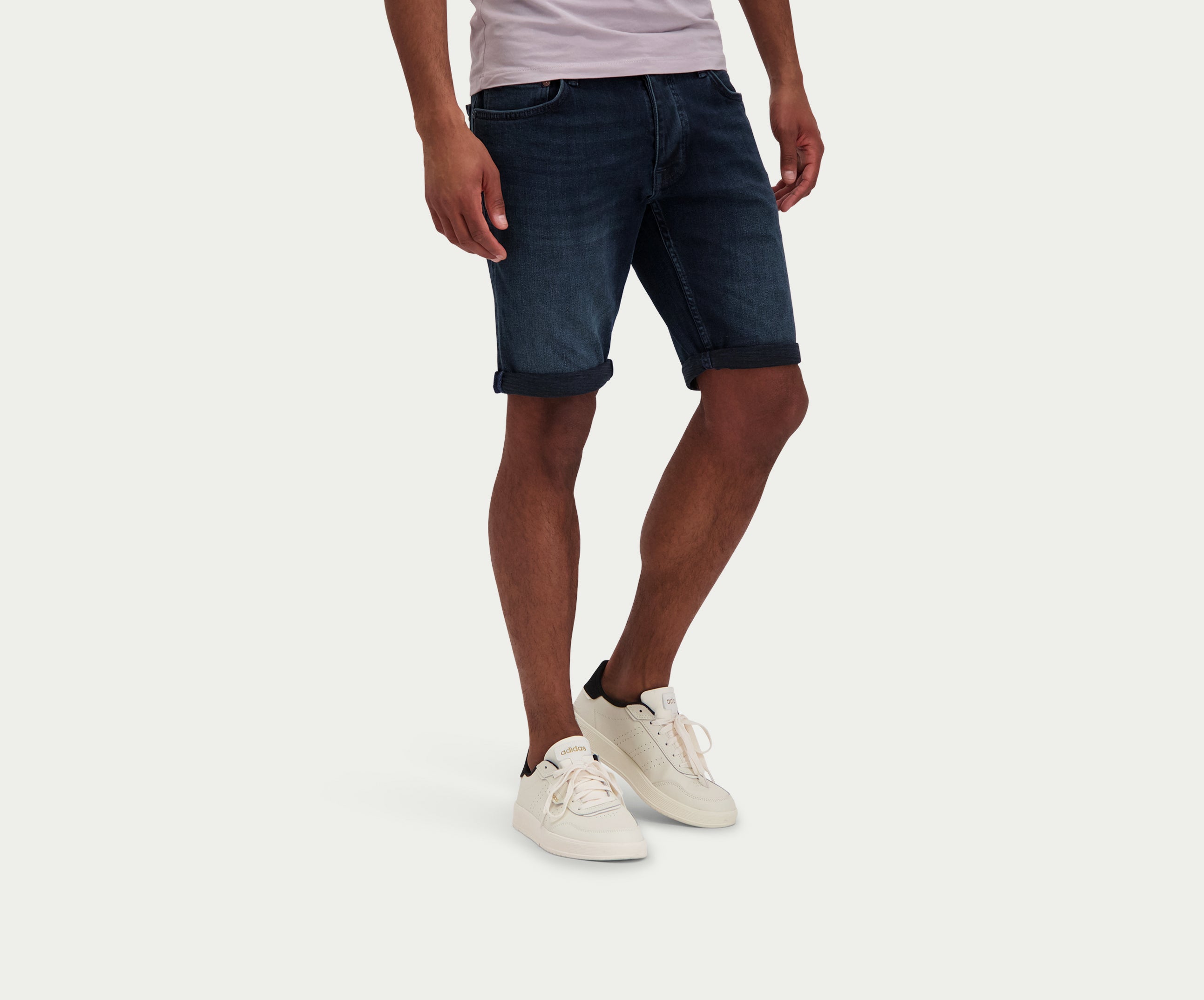 Een persoon die The N01 Short Blue Black van Blue Rivet Jeans draagt en witte sneakers staat tegen een effen witte achtergrond. Alleen de onderkant van het lichaam is zichtbaar.