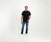 Een man staat tegen een effen witte achtergrond, draagt The N01 Tee Black, Blue Rivet Jeans en witte sneakers. Hij glimlacht met één hand in zijn zak, en straalt nonchalant zelfvertrouwen uit.