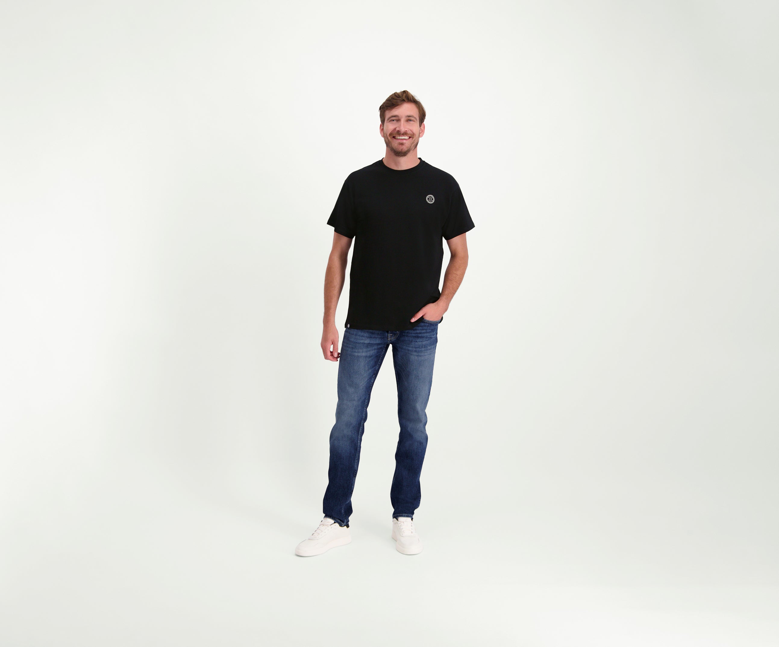 Een man staat tegen een effen witte achtergrond, draagt The N01 Tee Black, Blue Rivet Jeans en witte sneakers. Hij glimlacht met één hand in zijn zak, en straalt nonchalant zelfvertrouwen uit.