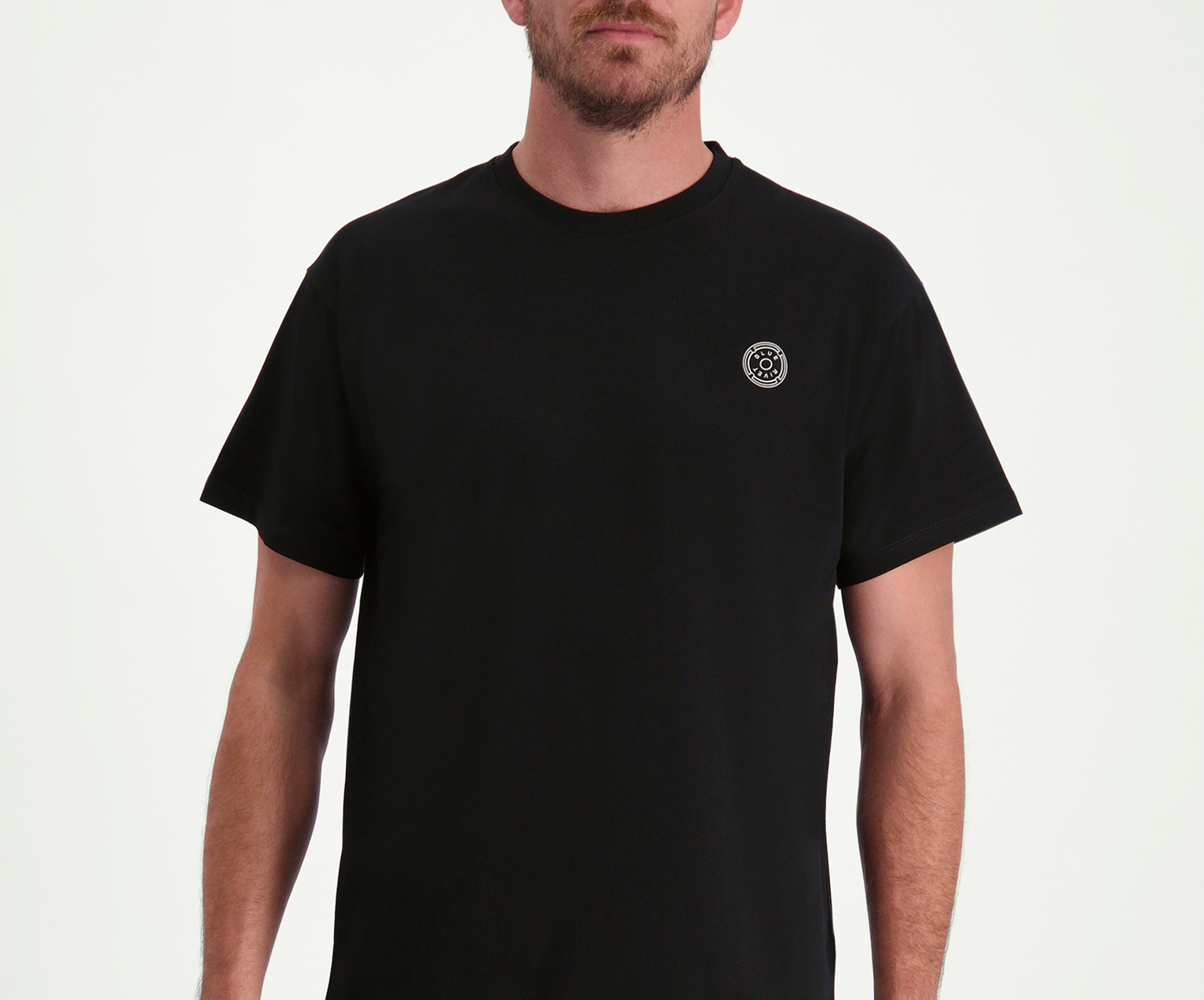 Een persoon die The N01 Tee Black van Blue Rivet Jeans draagt, gekenmerkt door zijn normale pasvorm en een subtiel klein rond embleem op de linkerborst, staat tegen een effen witte achtergrond. Het gezicht van de persoon is niet zichtbaar op de afbeelding.
