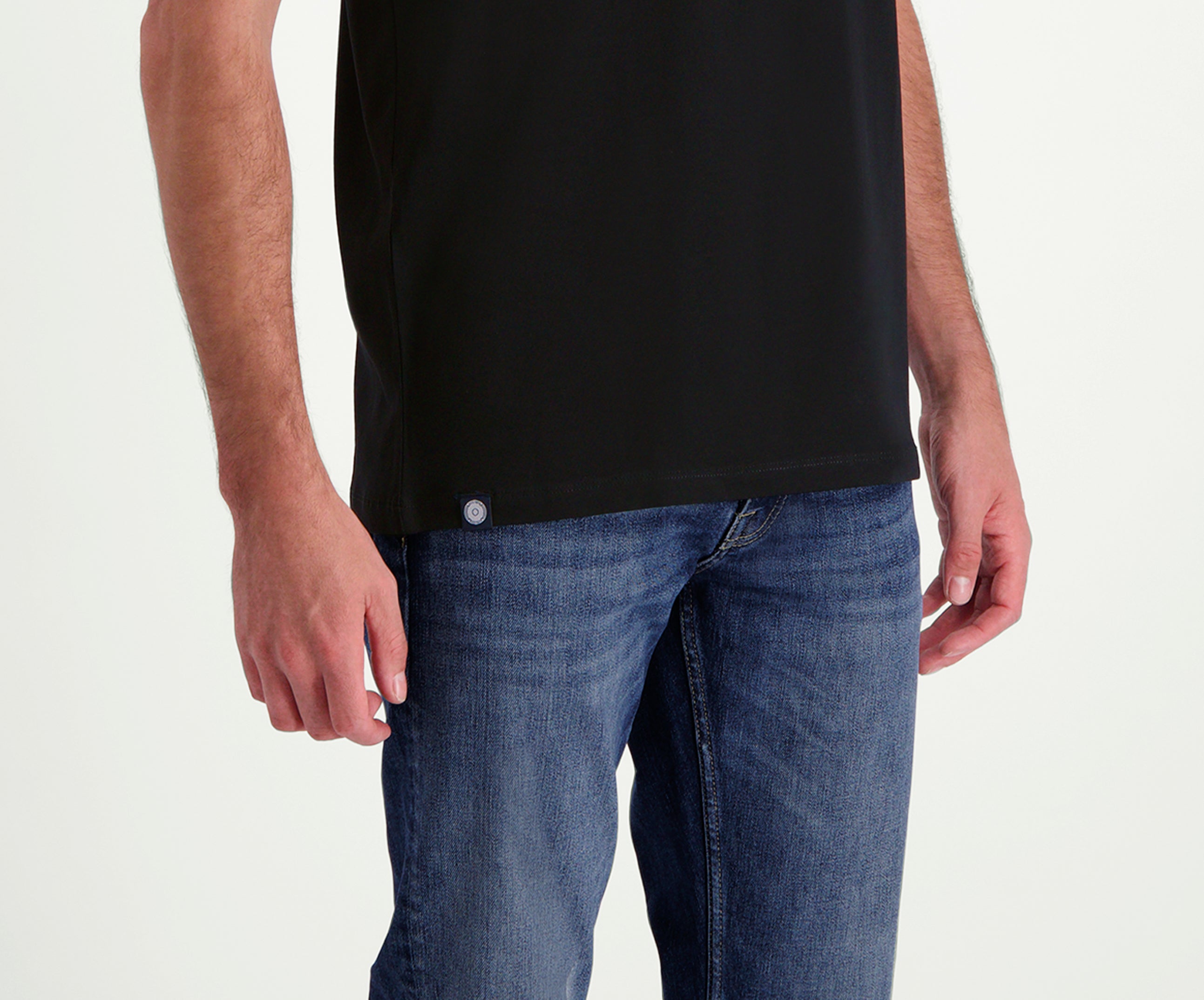Een persoon is afgebeeld vanaf de schouders naar beneden, met The N01 Tee Black in een normale pasvorm, gecombineerd met Blue Rivet Jeans. Beide armen zijn ontspannen langs hun zijden, met handen zichtbaar bij de heupen tegen een effen, lichtgekleurde achtergrond.