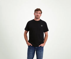 Een man met kort haar en een baard draagt The N01 Tee Black, een zwart regular fit Heren T-shirt met een klein rond logo op de borst, samen met Blue Rivet Jeans. Hij staat tegen een effen lichte achtergrond, handen in de zakken, en ziet er ontspannen uit.