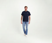 Een man draagt The N01 Tee Navy van Heren, lichtblauwe Blue Rivet jeans en regular-fit witte sneakers en staat tegen een effen witte achtergrond. Hij kijkt recht in de camera met een neutrale uitdrukking.